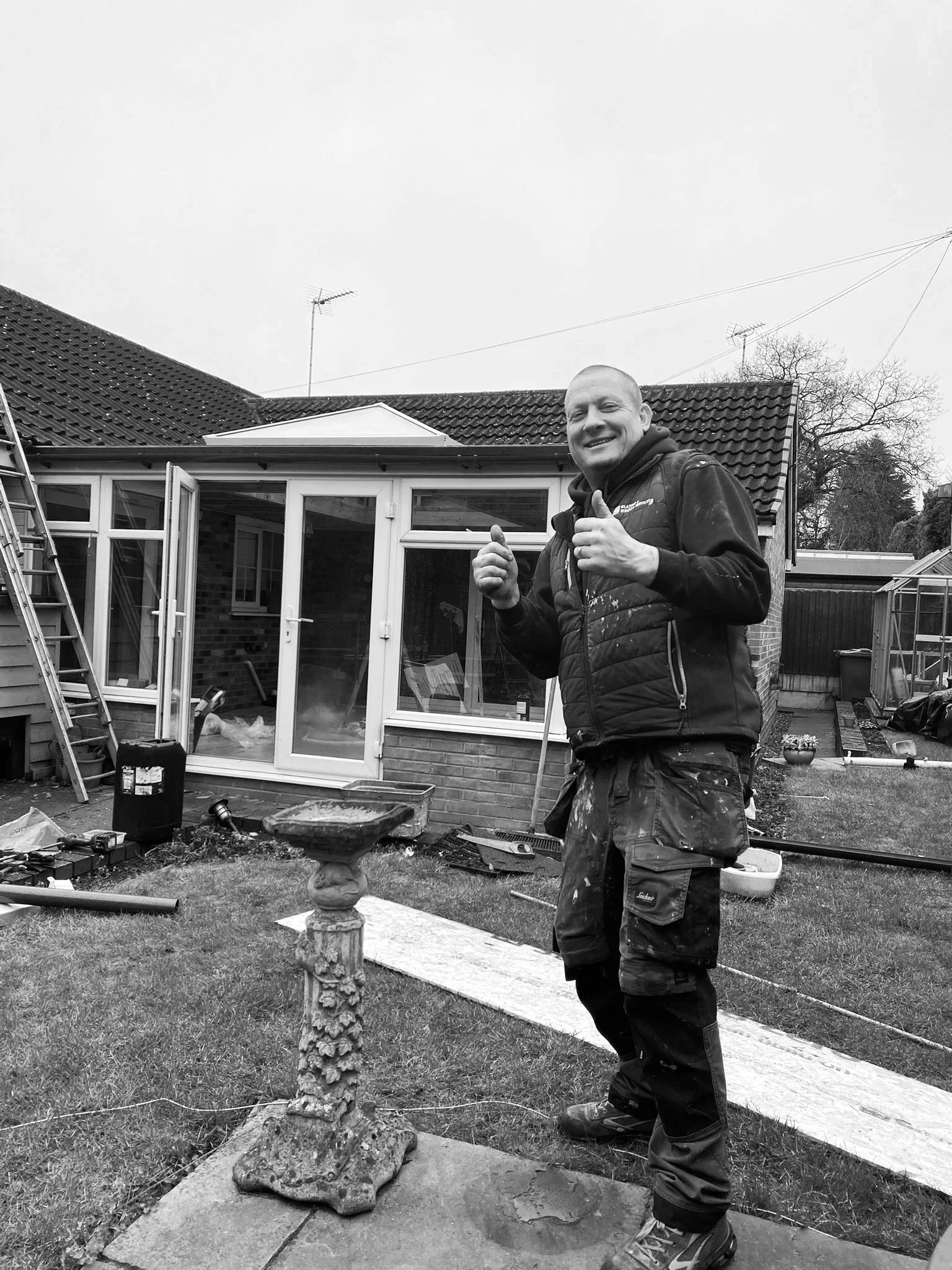 A smiling man in work clothes standing in a backyard, giving a thumbs-up gesture. There is a birdbath in front of him, and a house with a conservatory and open door behind him. Various tools and construction equipment are scattered around.