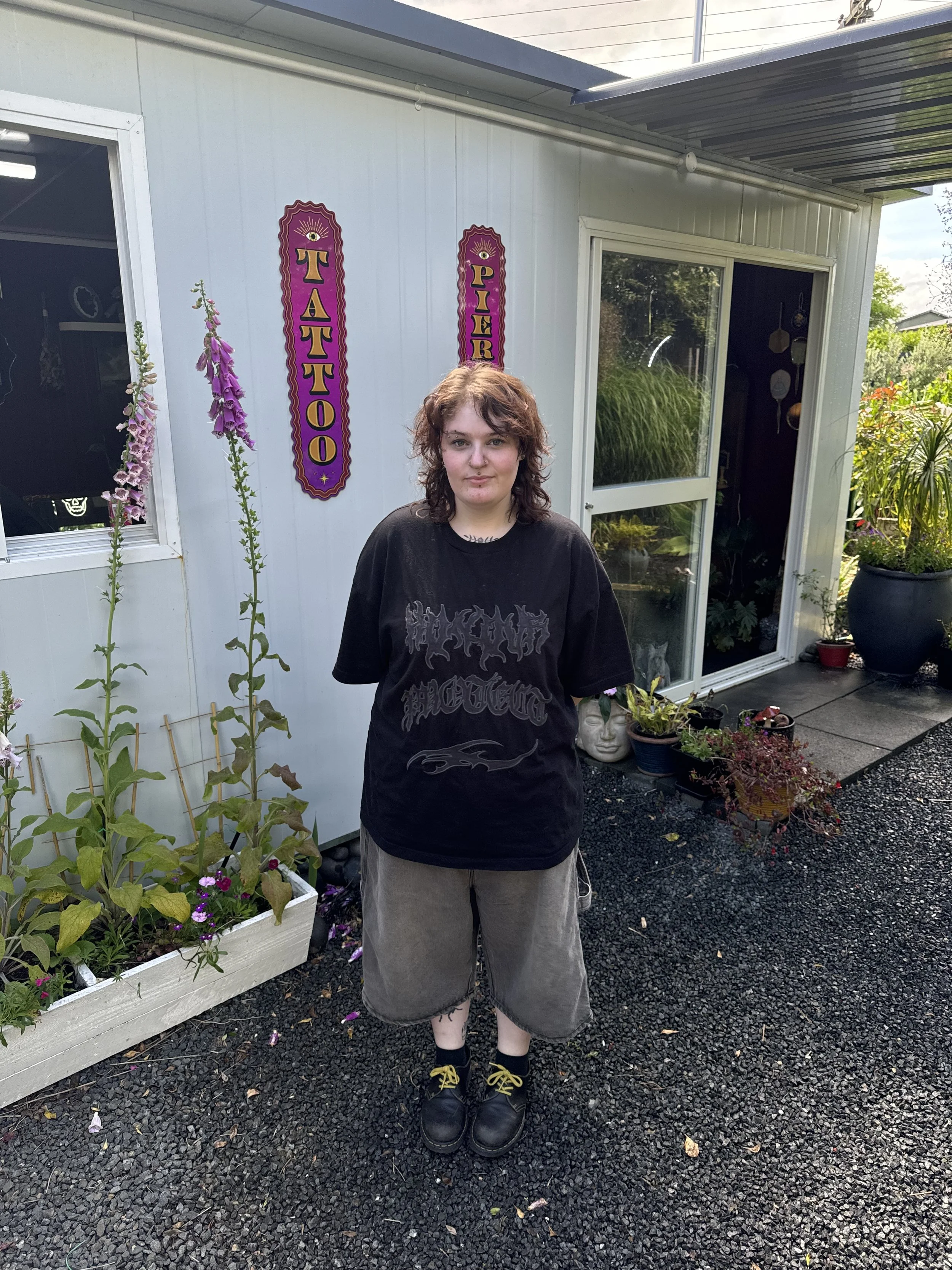 A person with curly brown hair standing outside near a building with signs that say "Tattoo" and "Pier". They are wearing a black graphic T-shirt, baggy shorts, black shoes with yellow laces, and black socks. There are pink and purple flowers and various potted plants nearby.