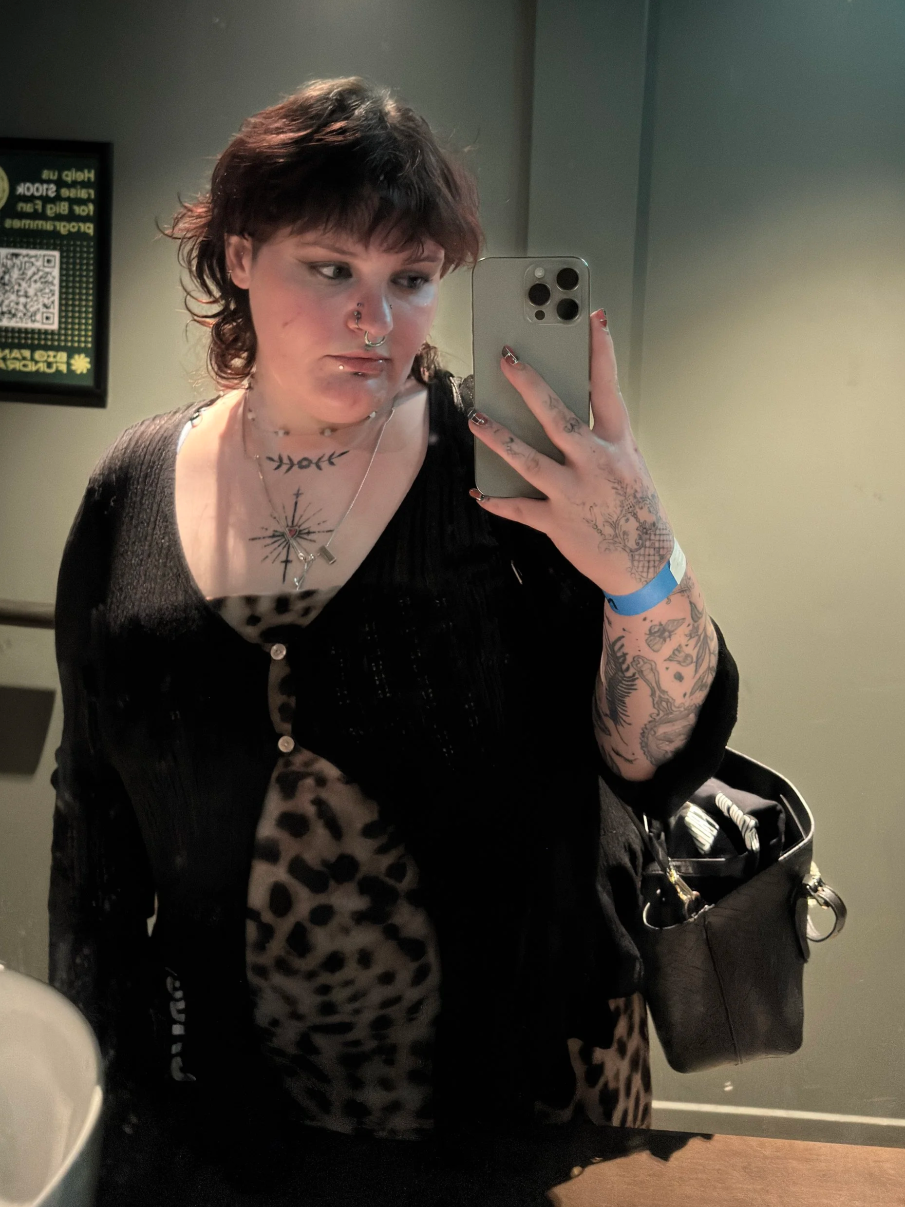 A woman taking a mirror selfie shows short, wavy dark hair with reddish highlights, multiple tattoos on her arms and chest, and facial piercings. She is wearing a black cardigan over a leopard print top and has a black bag hanging from her shoulder. She is holding a smartphone with a silver case in her right hand and is in a room with a gray wall and a green sign in the background.