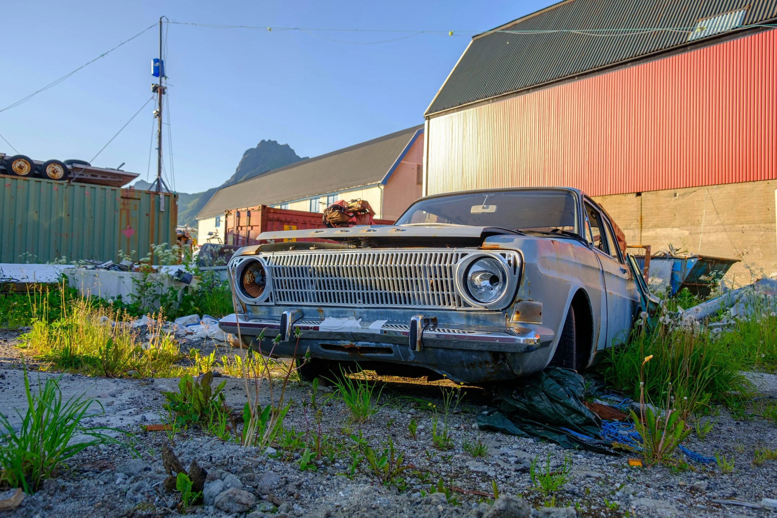 Old, abandoned car in a grassy and gravelly lot with colorful storage buildings and mountains in the background.