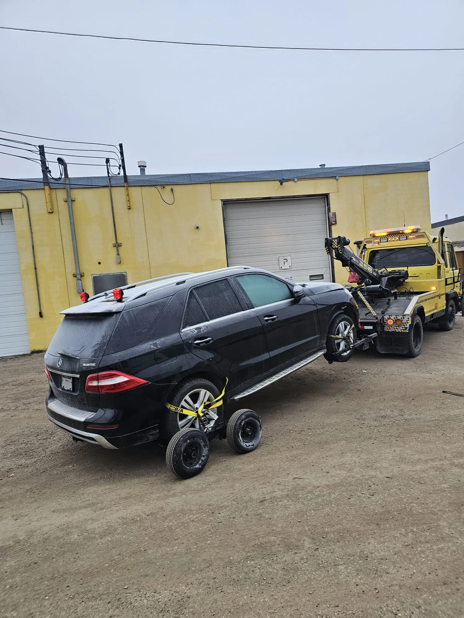 A black Mercedes-Benz SUV being loaded onto a yellow tow truck, with the SUV secured by yellow straps and the tow truck equipped with a hydraulic lift and hook. The scene is outdoors in front of a yellow building with multiple garage doors on a cloudy day.