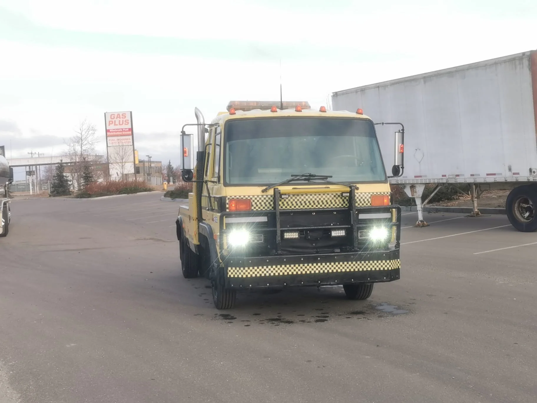 A yellow tow truck with flashing headlights in a parking lot, with trucks and a gas station sign in the background.