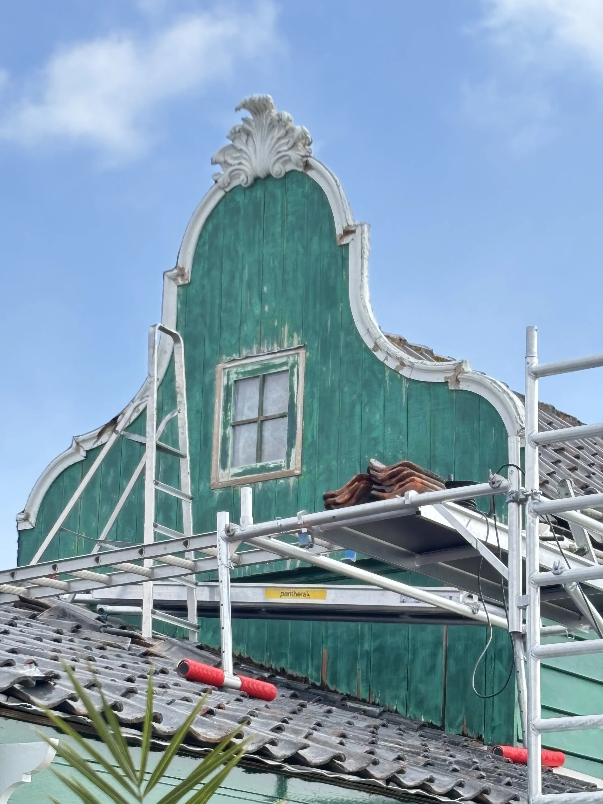 The image shows a building with classic Dutch-style gable, painted green, with a small window and white decorative trim. Scaffolding surrounds the building, indicating renovation or maintenance work.