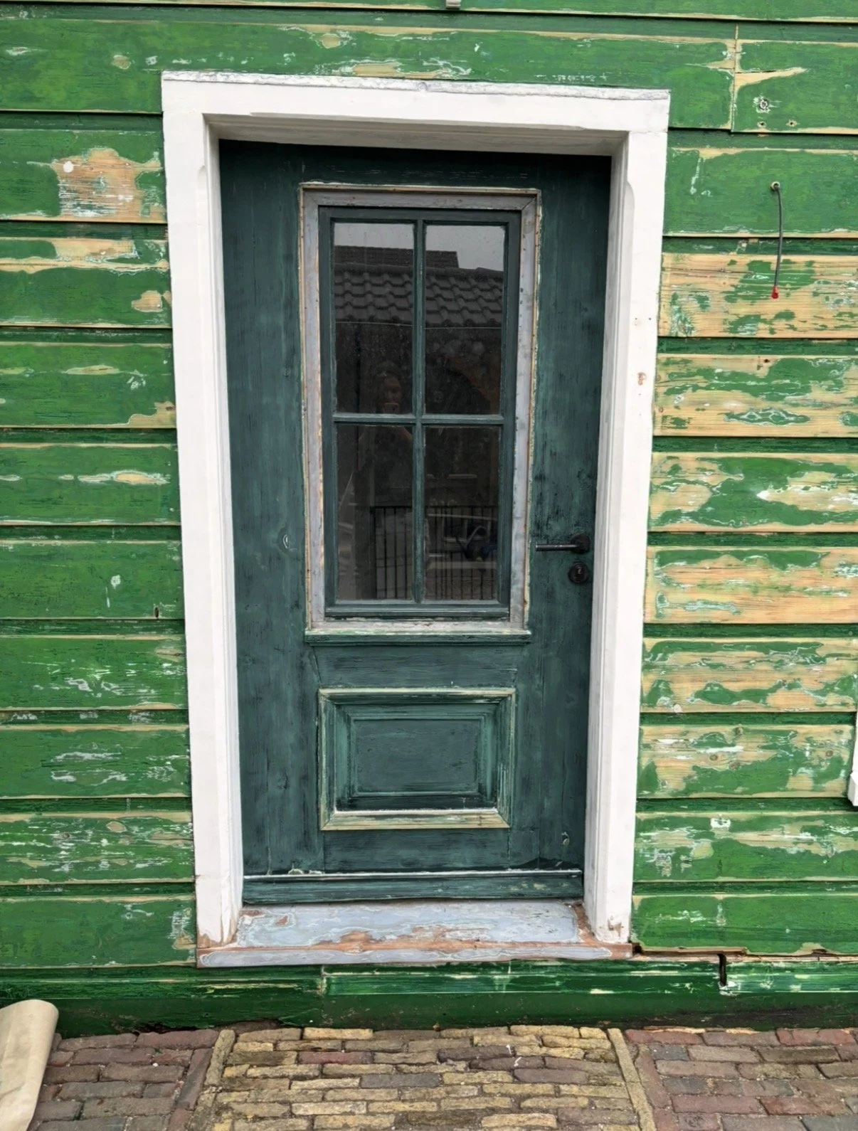 Groene houten deur met glas-in-lood ramen en witte lijst, behangen met verwerkte, geschilderde houten planken, op een stenen oprit.