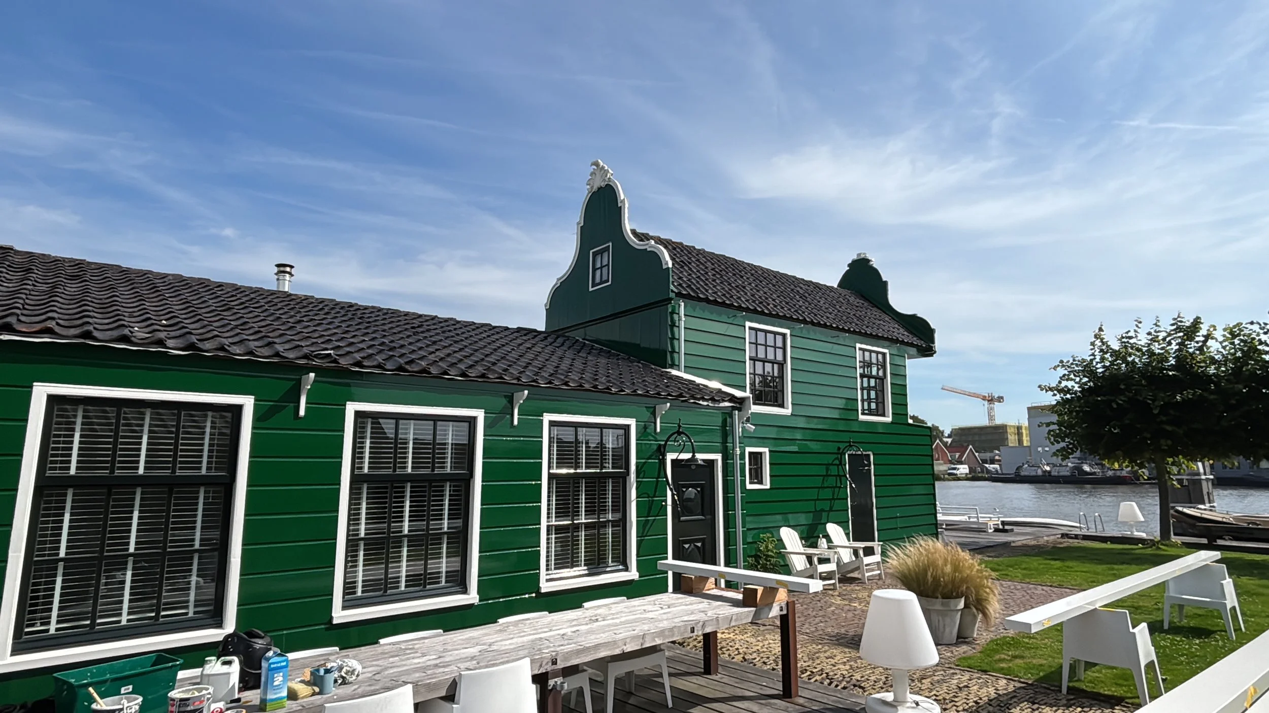 Groen huis aan de waterkant met witte ramen, buitenmeubilair, een boom, en een rivier met boten op de achtergrond onder een heldere hemel.