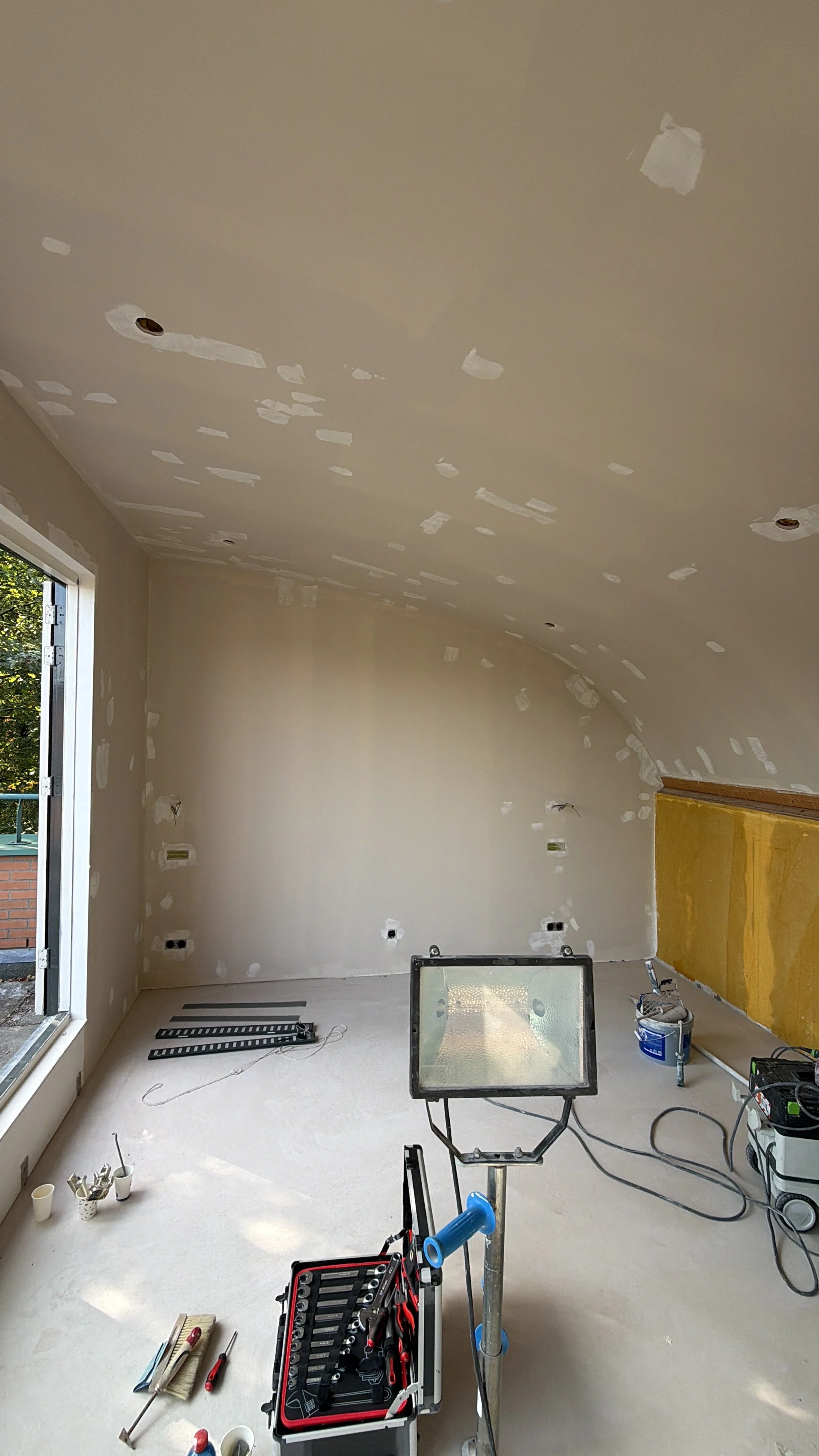 Interieur van een kamer in afwerking, halverwege renovatie, met spackwerk op de muren en het plafond, bouwgereedschap en materialen verspreid op de vloer, een grote lamp en een raam dat uit de muur is gesneden.