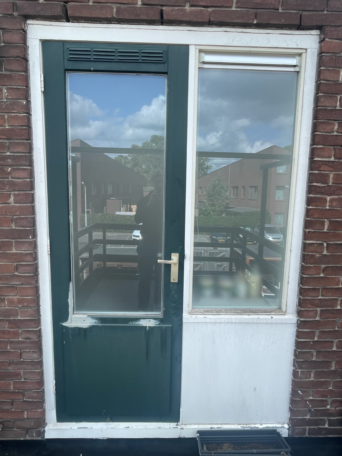 Buitenkant van een raam met een deur, die is vastgemaakt aan een bakstenen muur. Het raam bevat een grote glazen ruit die de weerspiegeling van de lucht en huizen toont.