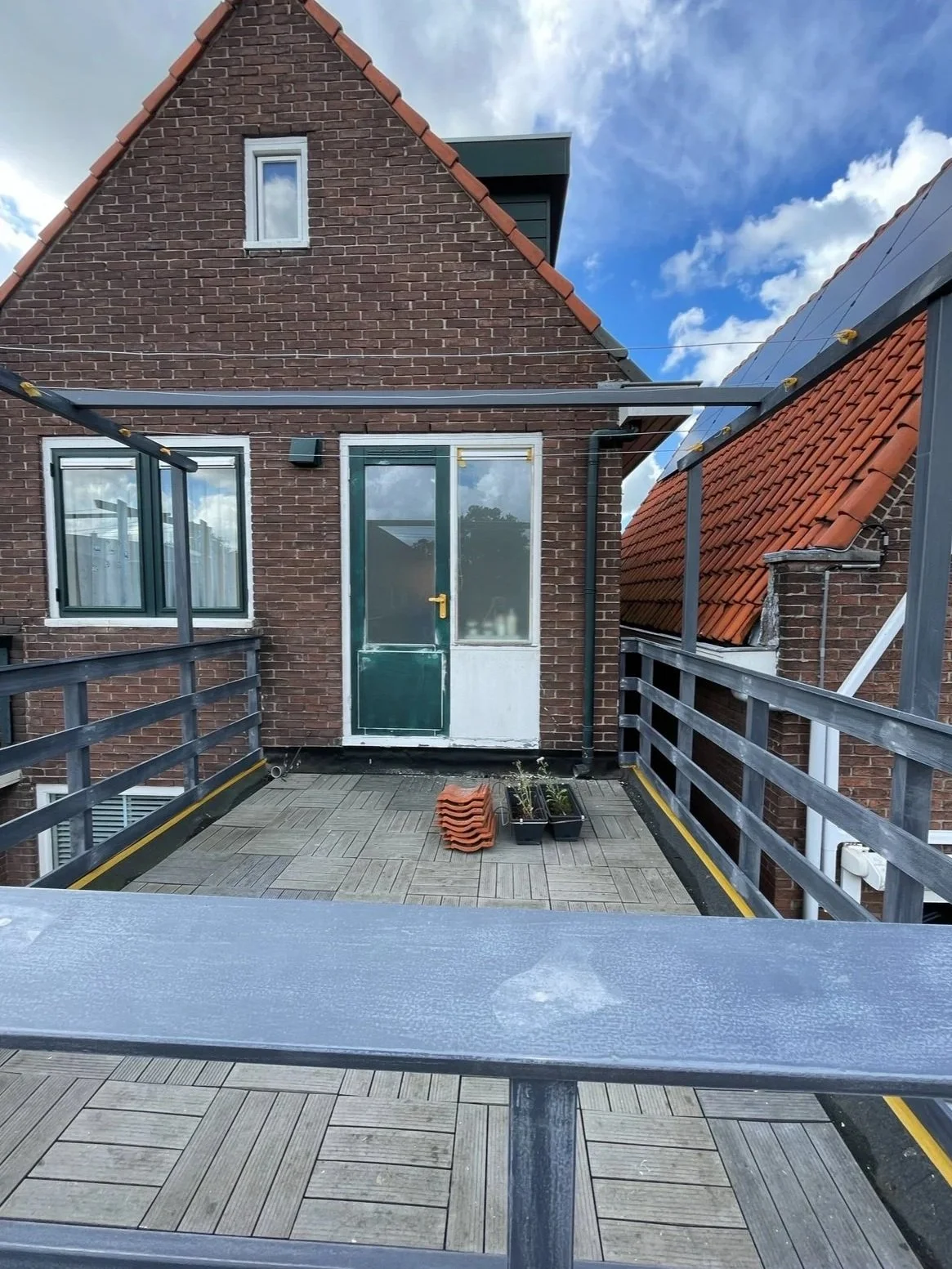 Balkon met houten vloer, plantenbakken met jonge planten, buitendeuren, ramen en een tuin van een woning met bakstenen muren en een variëteit aan dakpannen.