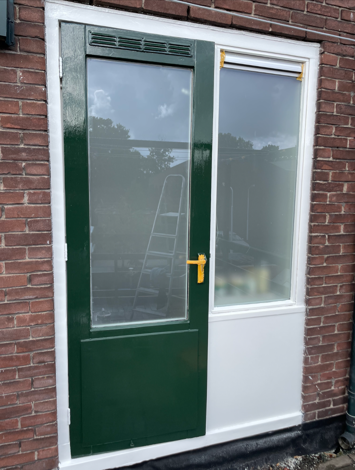 Roze deur en raam in een bakstenen muur, met een ladder zichtbaar in de weerspiegeling.