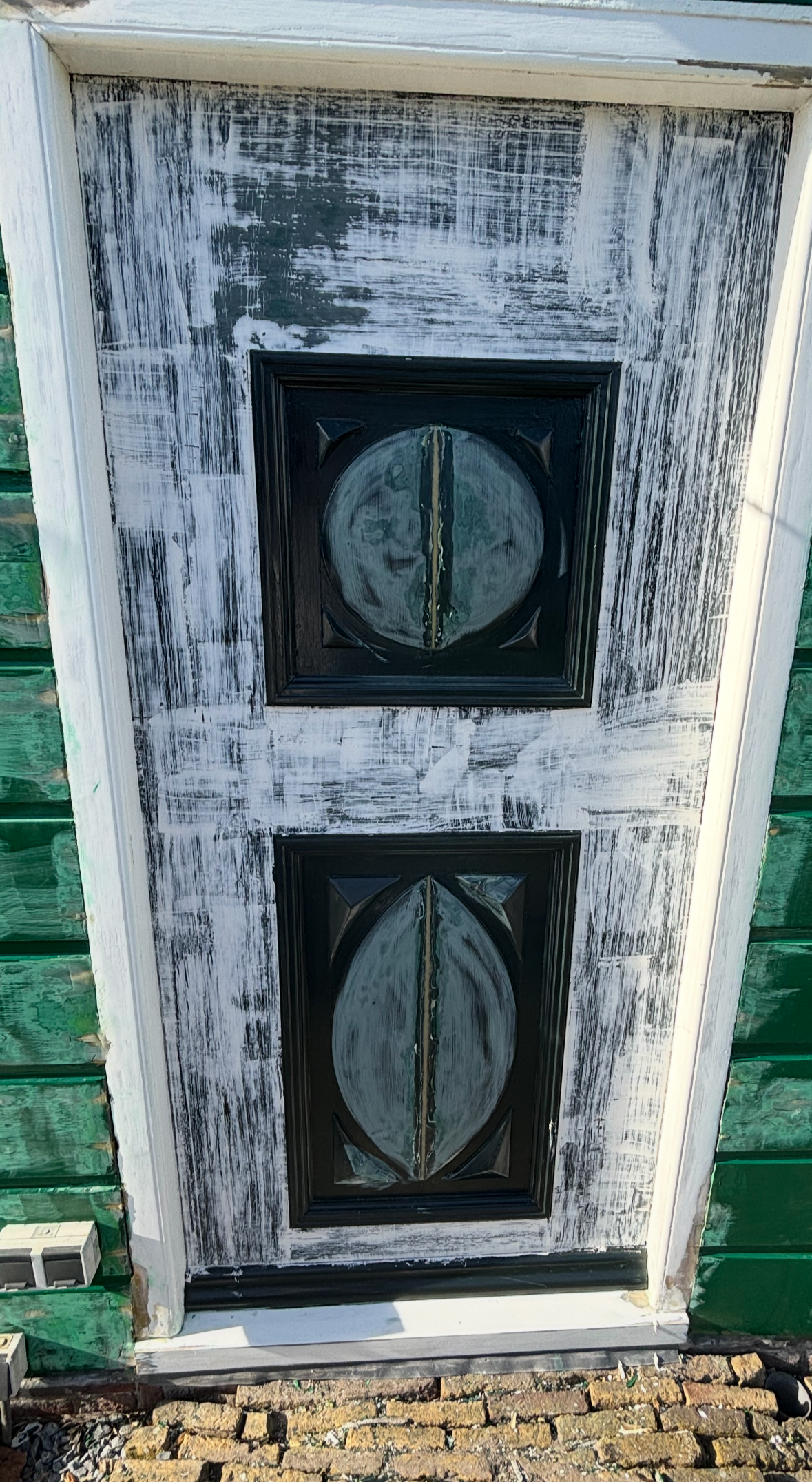 Buitenkant van een deur met een zwarte ruit en een houten frame dat geschilderd wordt, met groen verlichte muur ernaast.