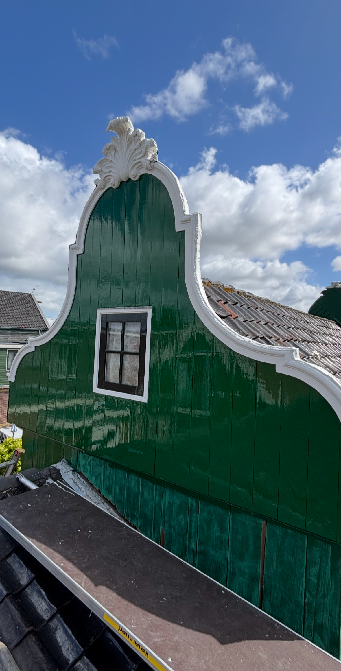 Een groene gevel met wit sierlijk lijntje en een klein zwart raam, onder een blauw met witte wolken hemel.