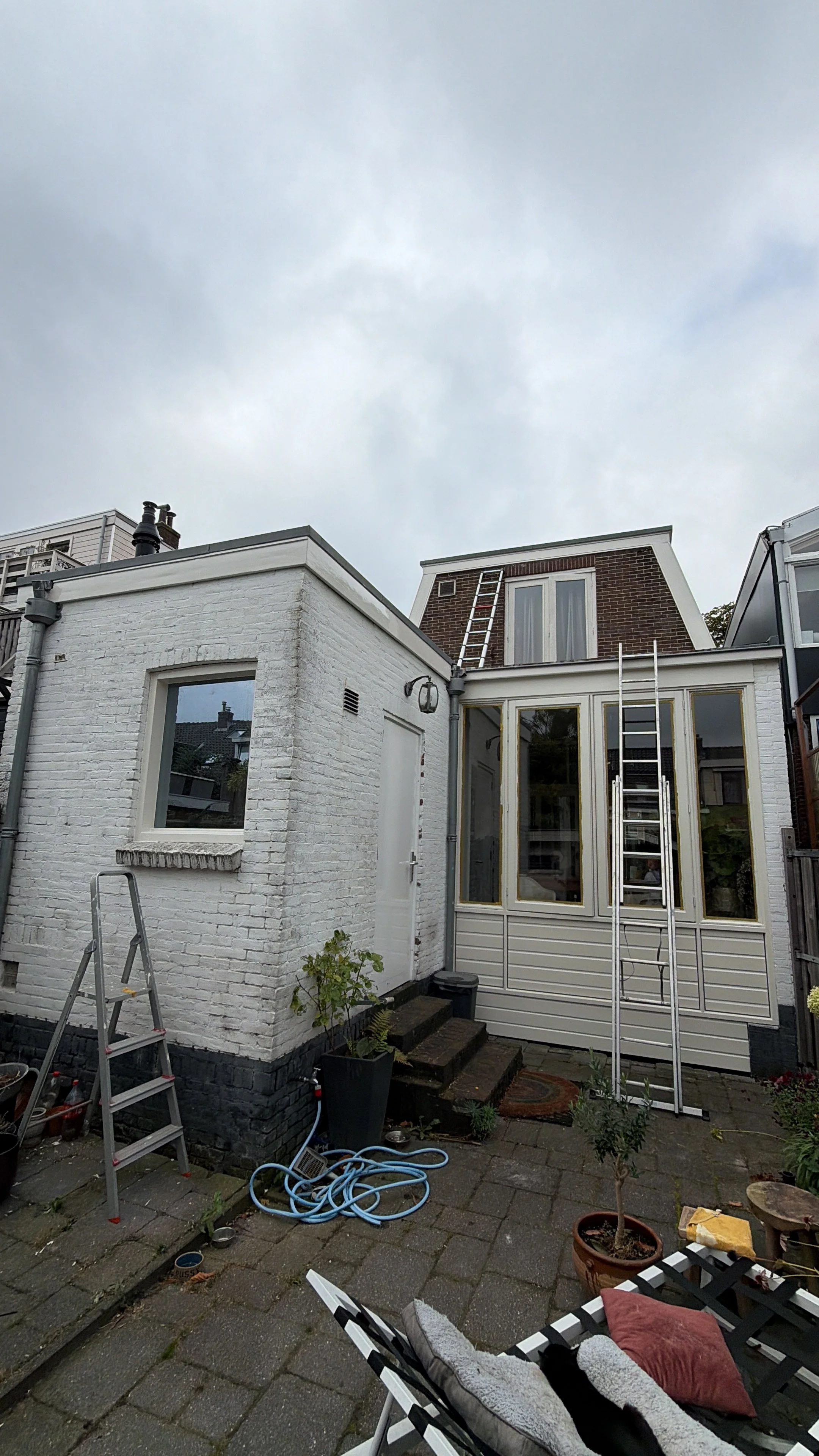 Twee gebouwen achter elkaar met ladders op het dak, een tuin met planten, tuinmeubilair en tuingereedschap, een blauwe tuinslang en een gemetseld pad.