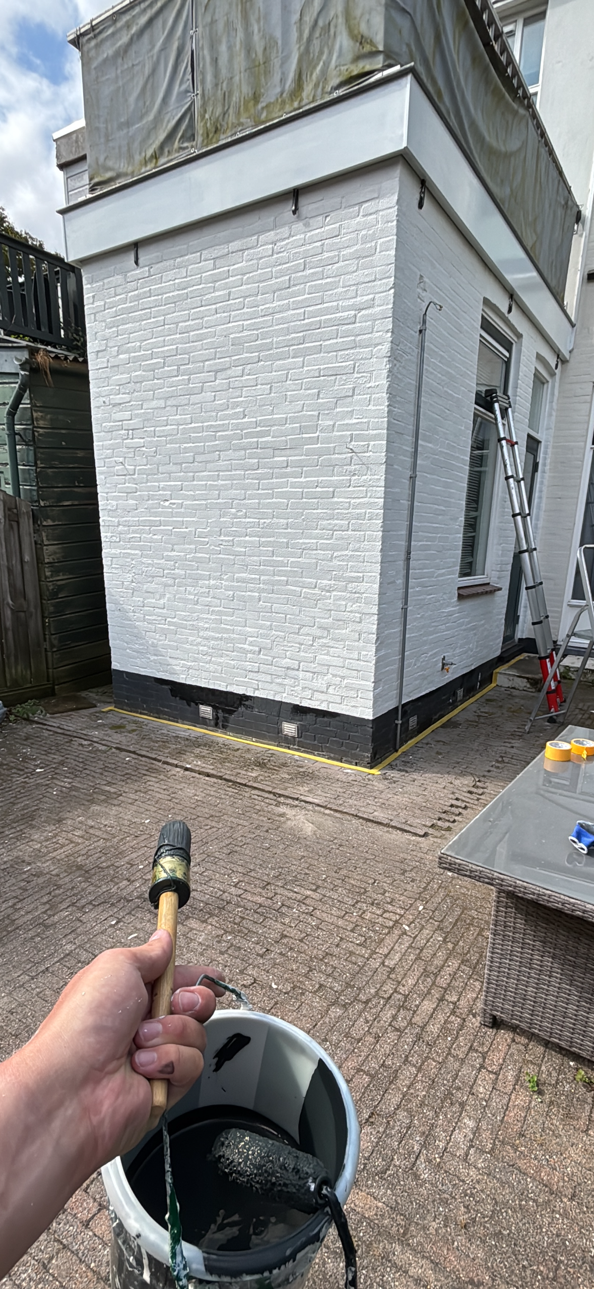 Een persoon die verf aanbrengt op een witte gevel van een gebouw met een verfroller en een bak verf, met een ladder en tuinmeubilair op de achtergrond.