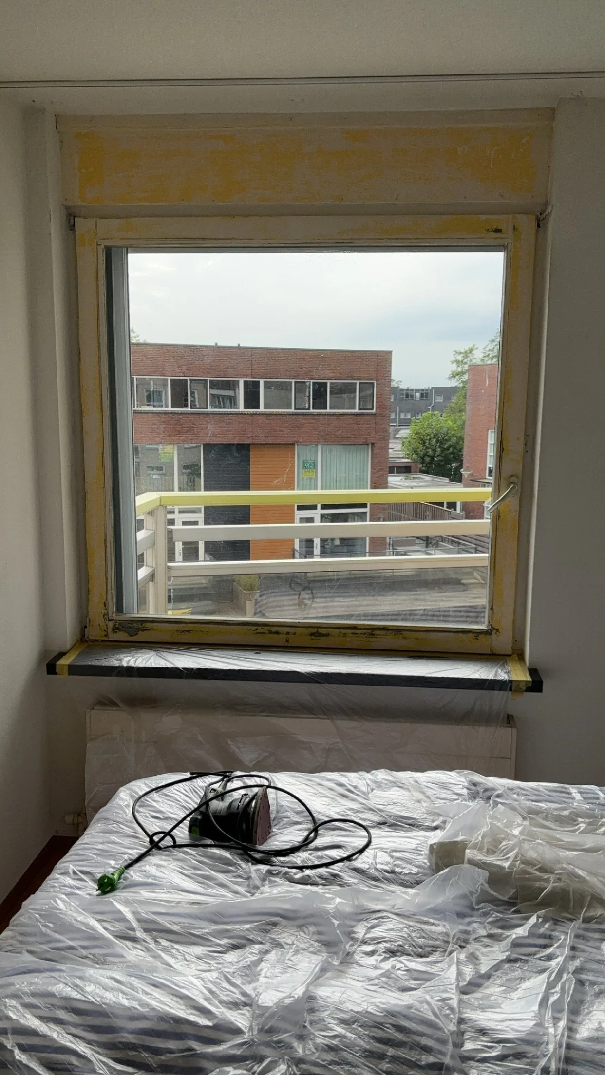 Binnenkant van een kamer met een in verval geraakte vensterkozijn en een bed dat bedekt is met beschermfolie, met een elektrische adapter en een stekker op het bed.