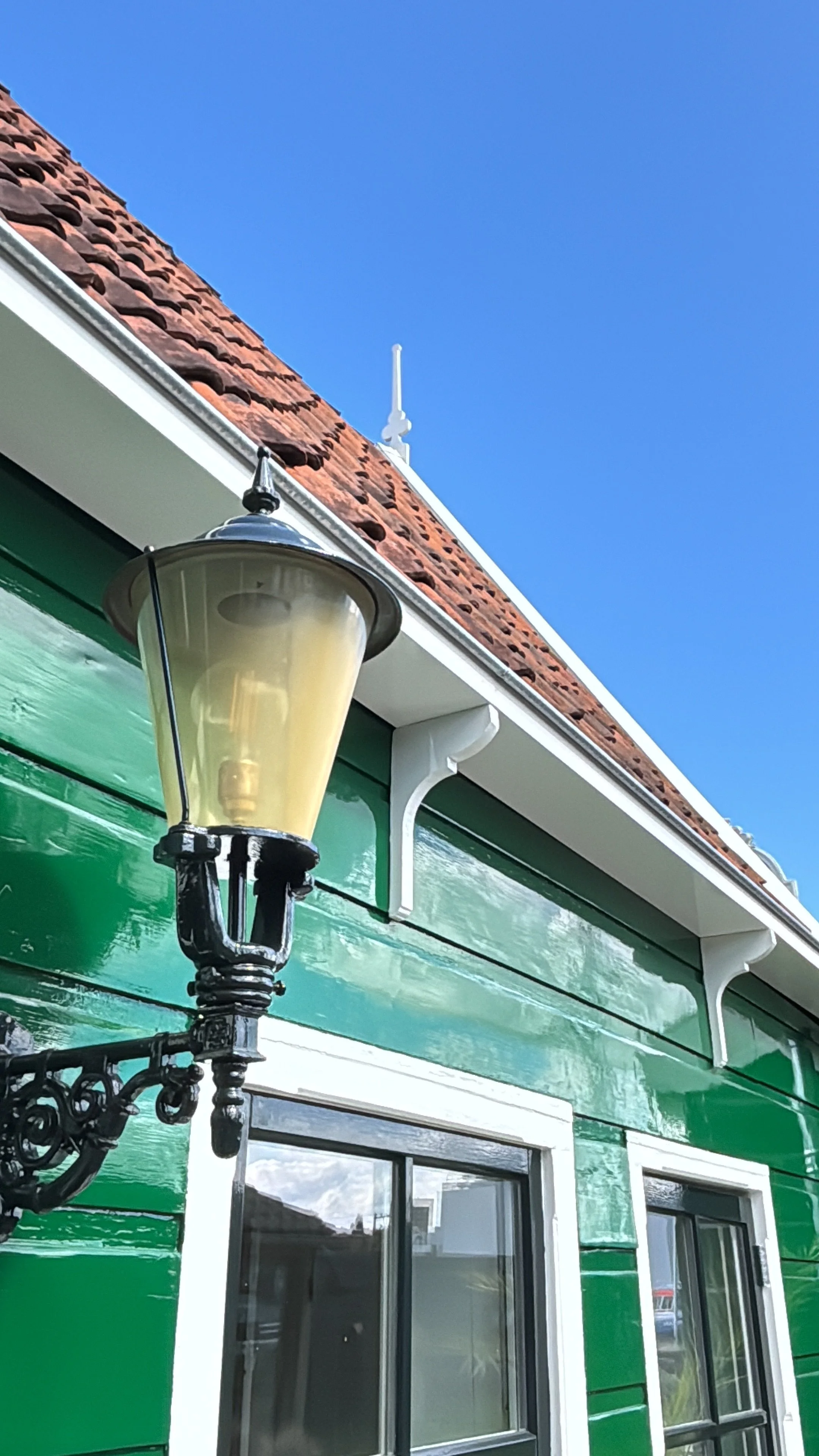 Groene houten gevel met witte raamkozijnen, een zwarte klassieke buitenlamp en een rood pannendak onder een blauwe hemel.