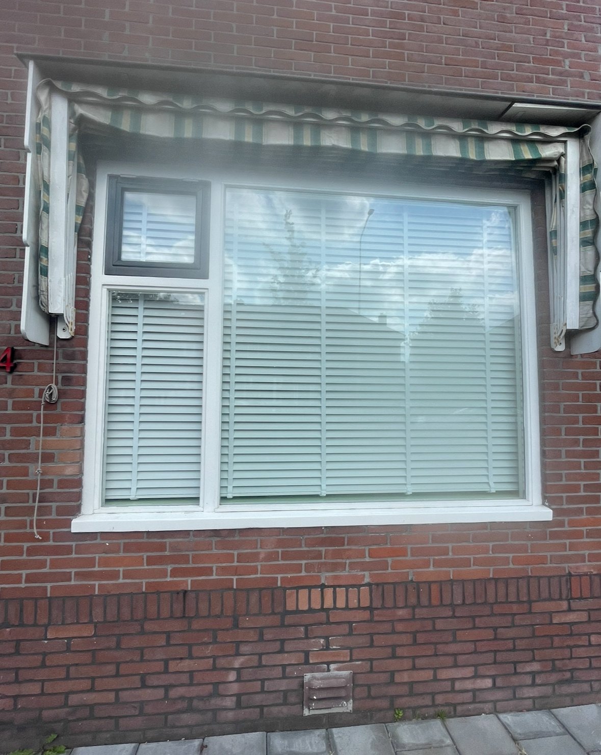 Een woonkamerraam met witte jaloezieën in een rode bakstenen muur, met een uitvalscherm erboven.