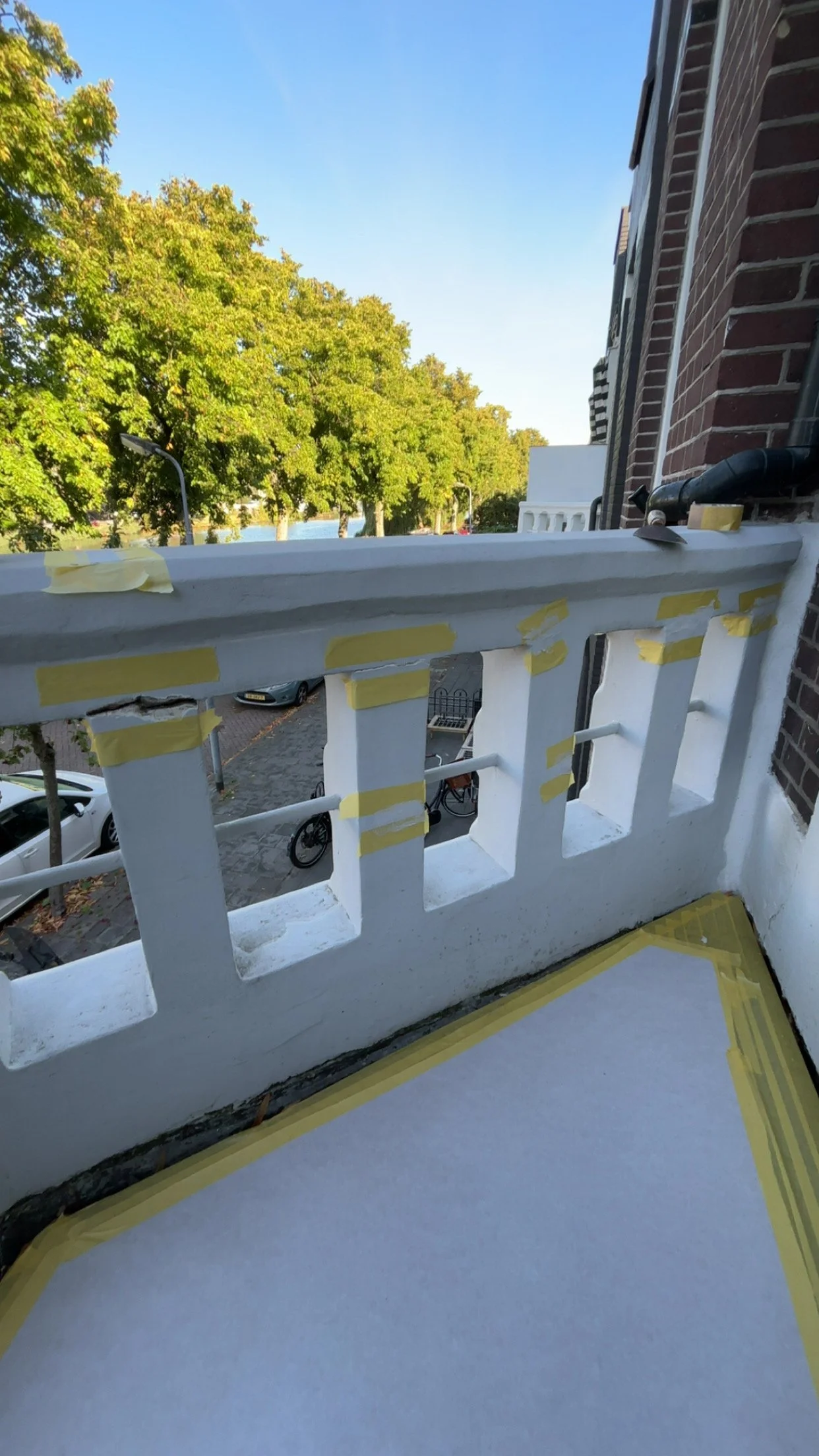 Kleine balkon met witte muur en gele tape, uitzicht op straat met bomen, auto’s en fiets, en een stukje huismuur.