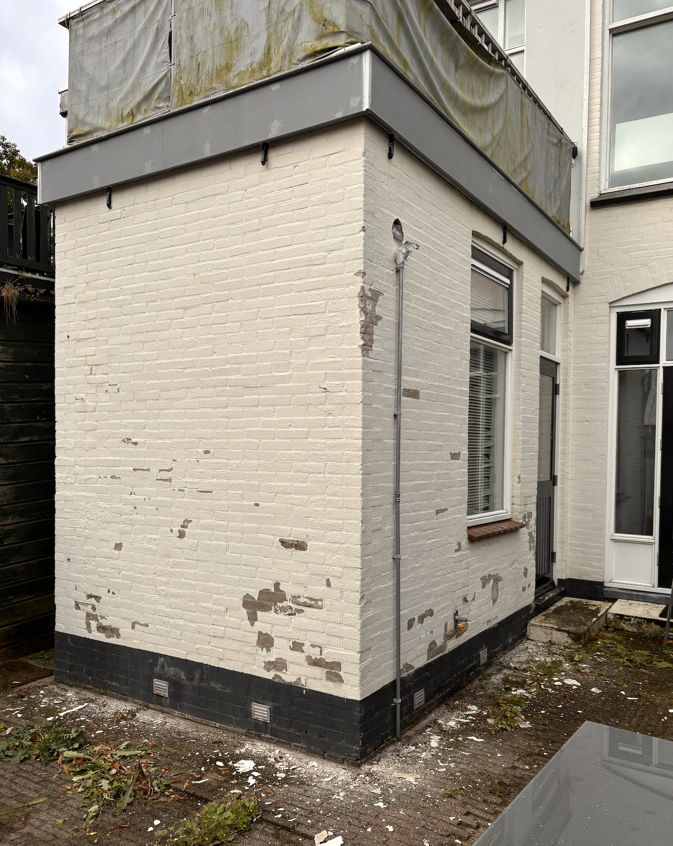 Buitenkant van een huis met witte bakstenen muur en enkele beschadigingen, een raam en een deur, een verongelukte regenpijp en een zwartgelakte voet van de muur.