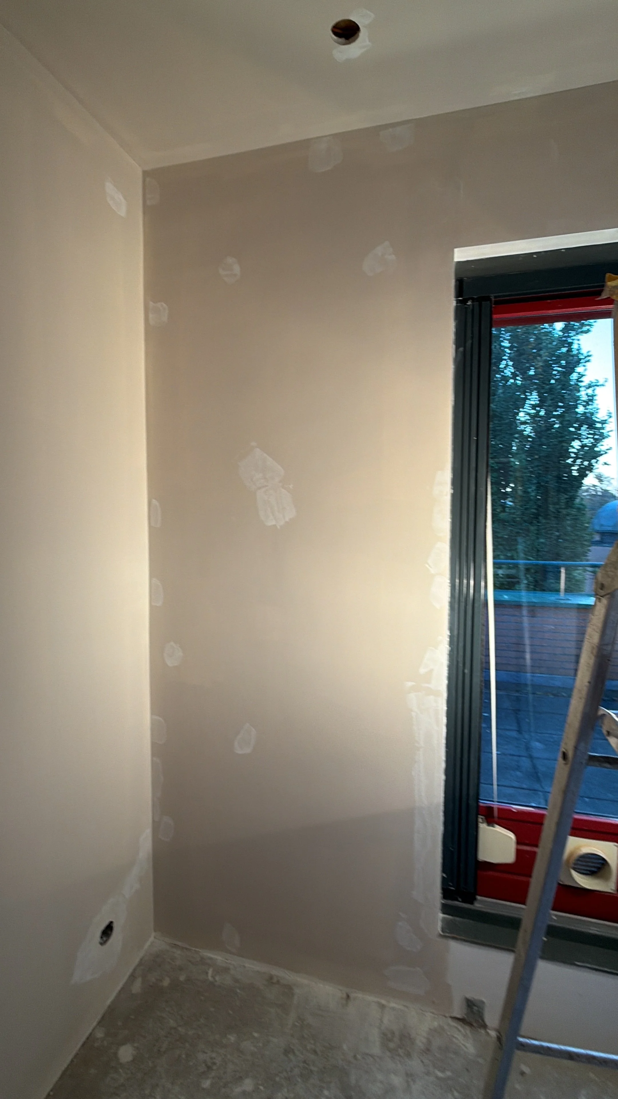 Binnenmuur in aanbouw met gipsplaten en schuurplekken, naast een raam met uitzicht op bomen, tijdens renovatie of bouw.