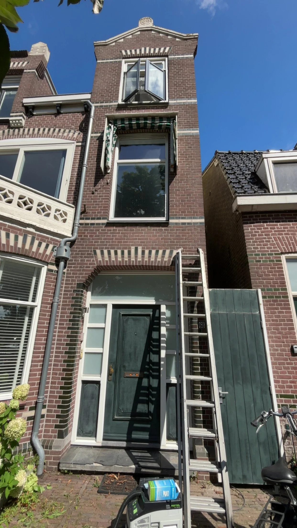 Een drieverdien huis met een groene deur, een ladder voor de deur, een emmer en een trap. Er staan ook een fiets en een machine voor het schoonmaken of klussen.