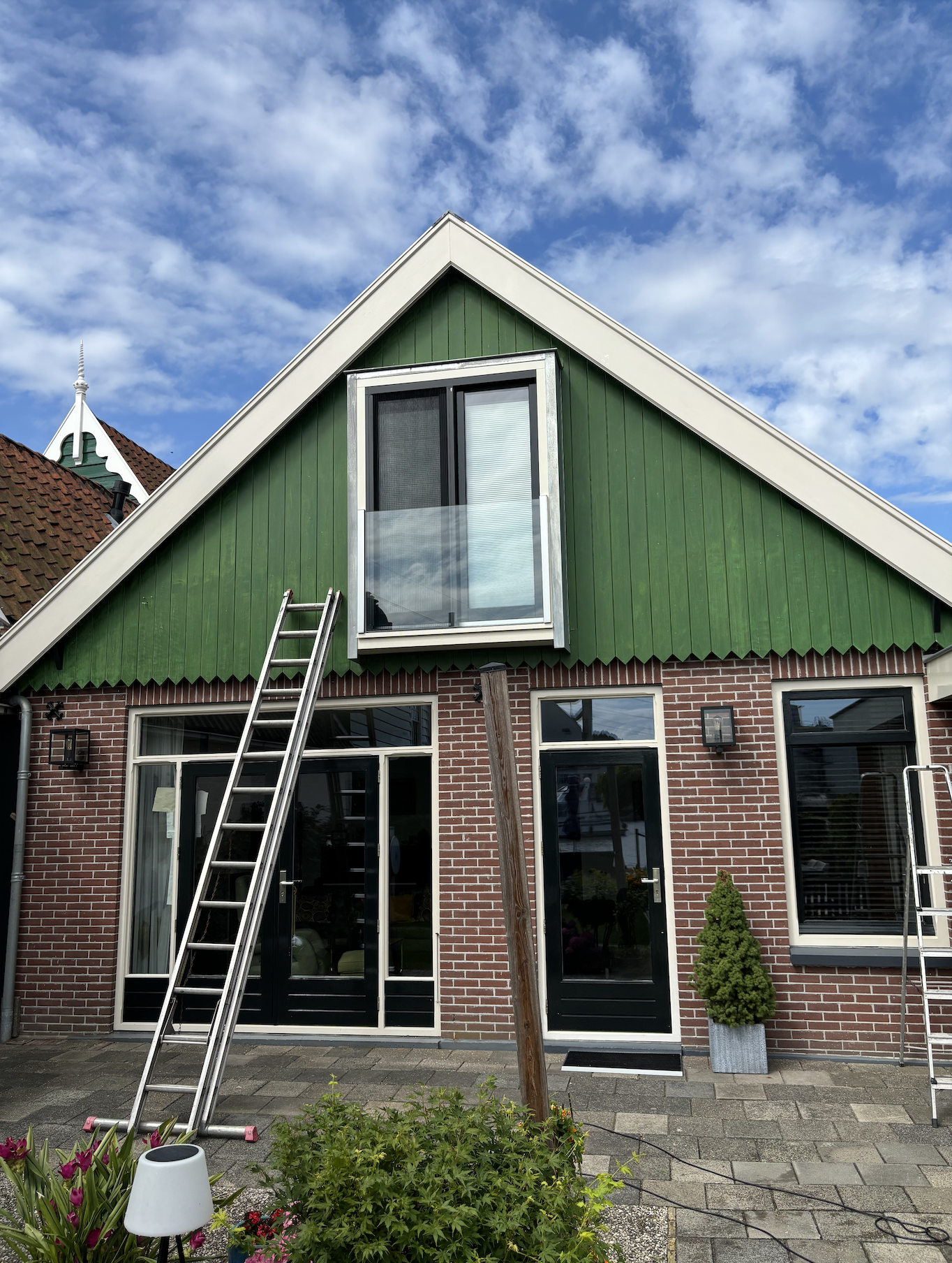 Een huis met een rode bakstenen gevel, grote ramen en een groene gevel boven, een ladder voor de gevel en een boom in een plantenbak. Een blauwe sky met wolken.