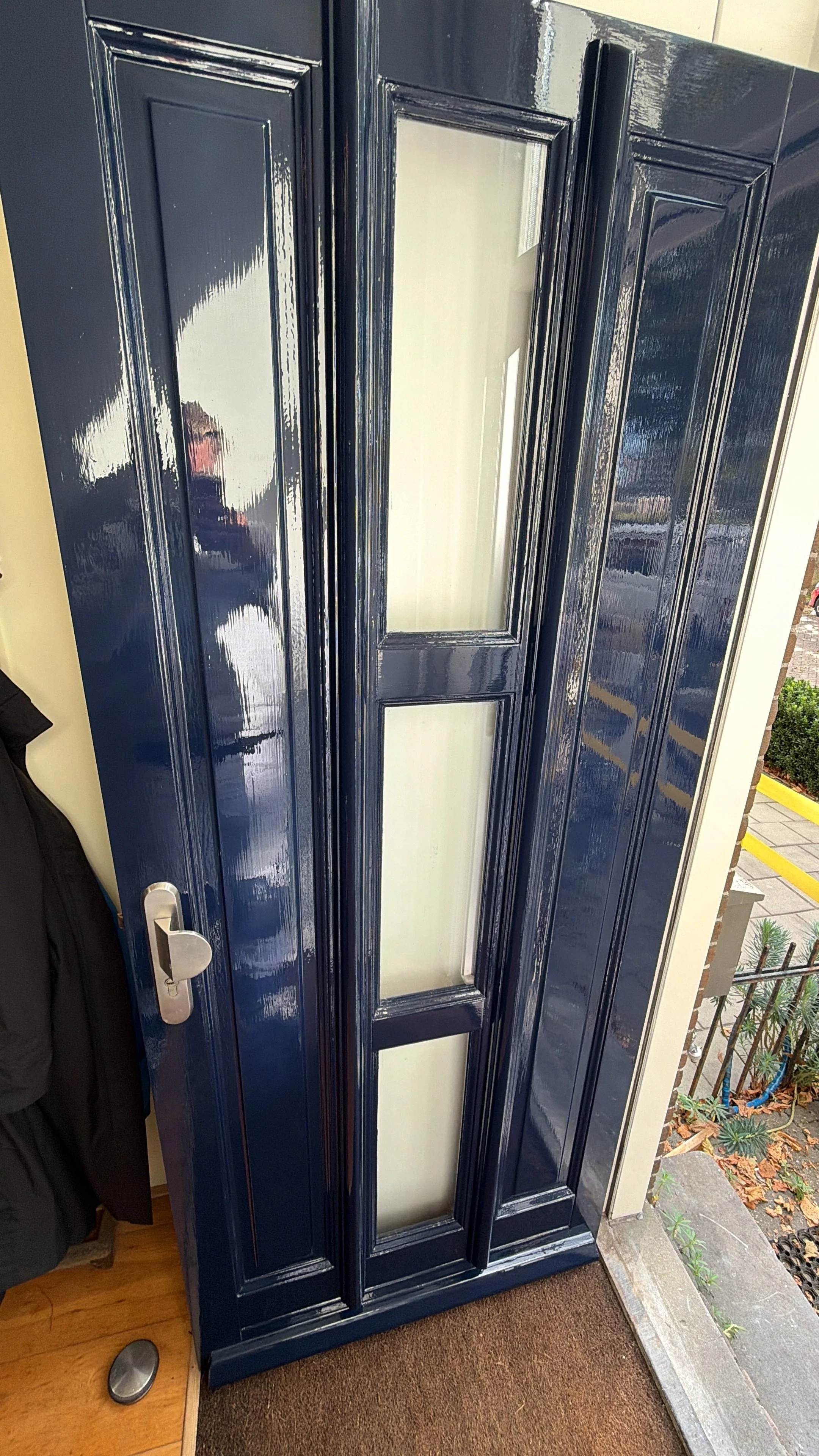 Een donkerblauwe voordeur met drie glazen panelen en een zilverkleurige deurknop, voor een woning met een betegelde stoep. Er is een buitenraam en planten te zien buiten.