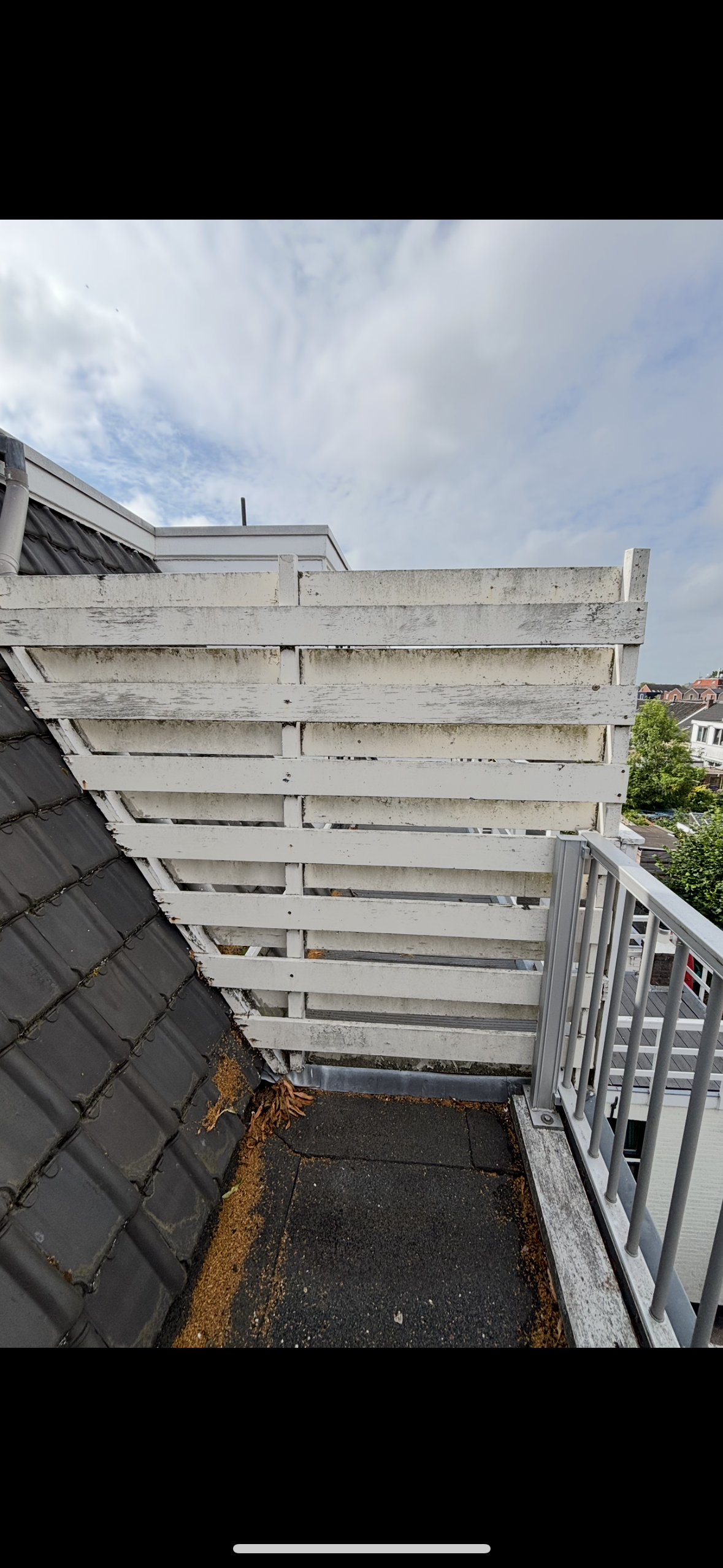 Een balkon met een witte houten afscheiding die vuil en schimmel vertoont, naast een kozijn met een hellend dak bedekt met zwarte dakpannen. Op de vloer liggen bladeren en vuil, met uitzicht op een buurt met bomen en huizen onder een bewolkte lucht.