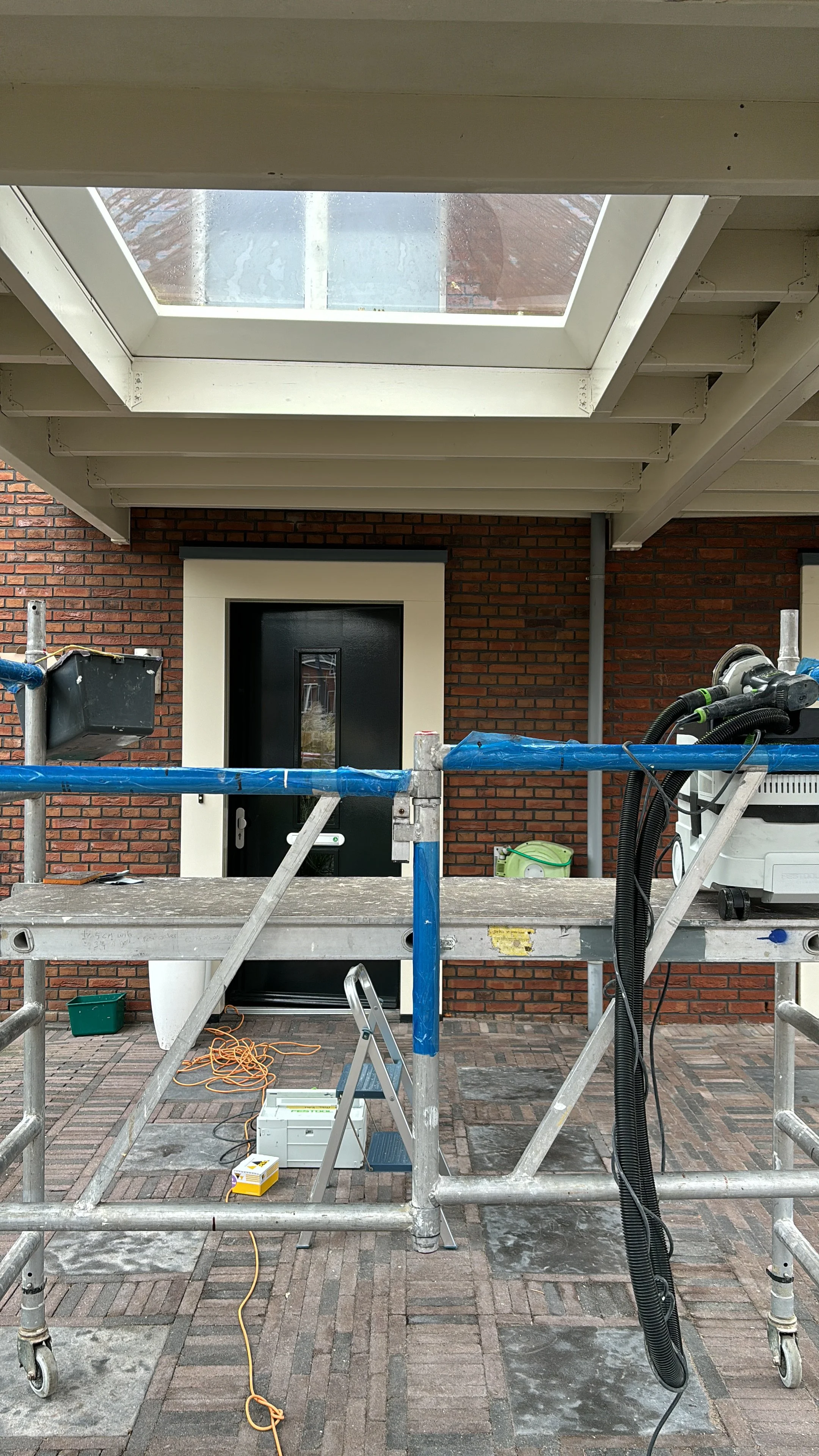 Bouwsteiger voor werkzaamheden aan de gevel van een huis, met een zwarte deur, bruin bakstenen muur, en een dakraam of lichtkoepel in het plafond.