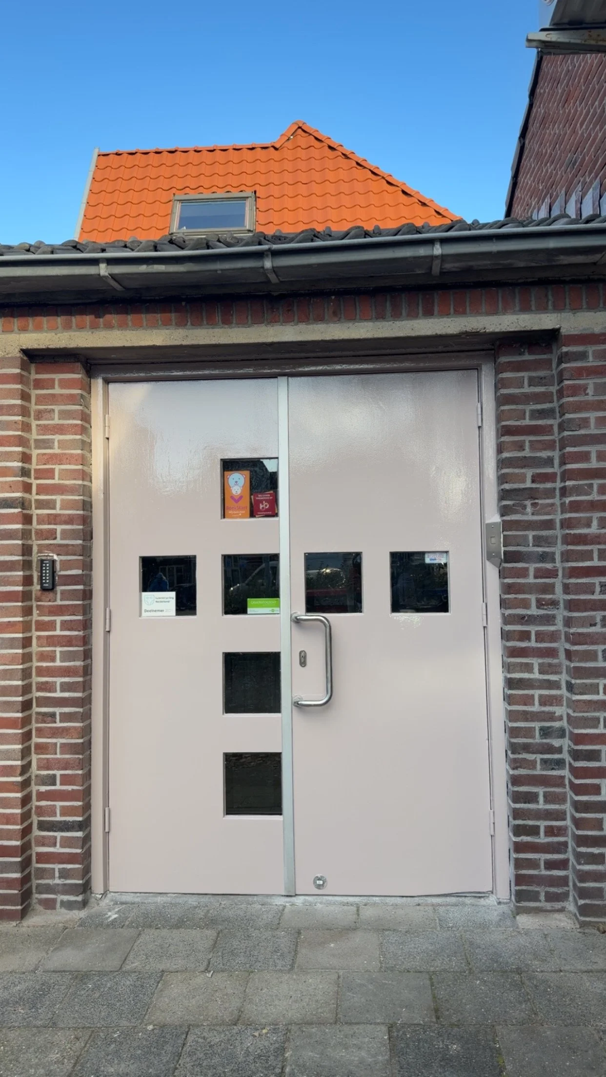 Dubbele toegangsdeur met kleine vierkante ramen, gevel van baksteen, rood dak met een schuin dak en een dakraam, in een tuin of binnenplaats onder een heldere blauwe lucht.