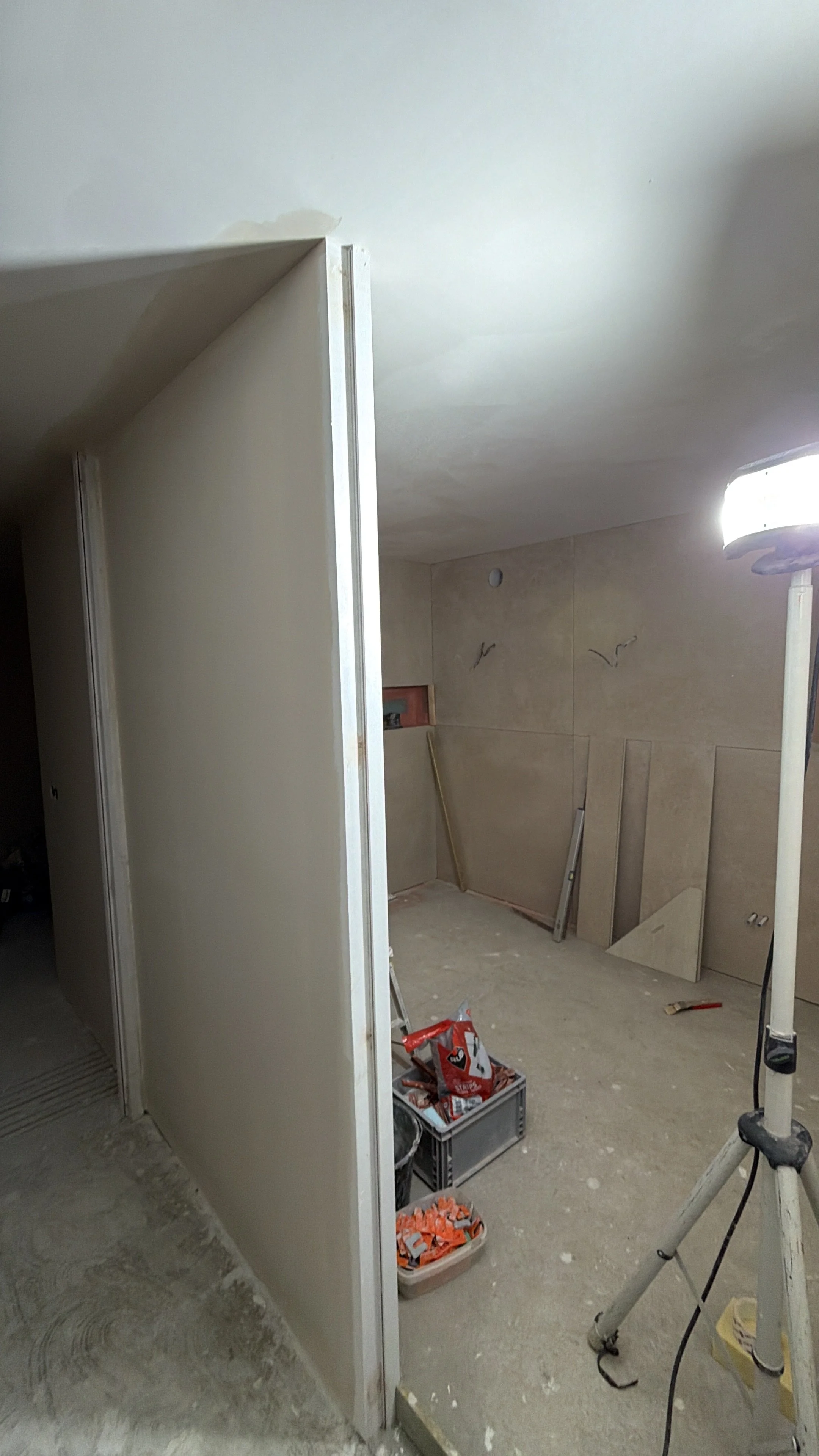 Een kamer onder constructie met bouwmaterialen en gereedschap. Er is een scheidingswand en een muur met bekleding, een werklamp en opslagbakken met bouwmateriaal.