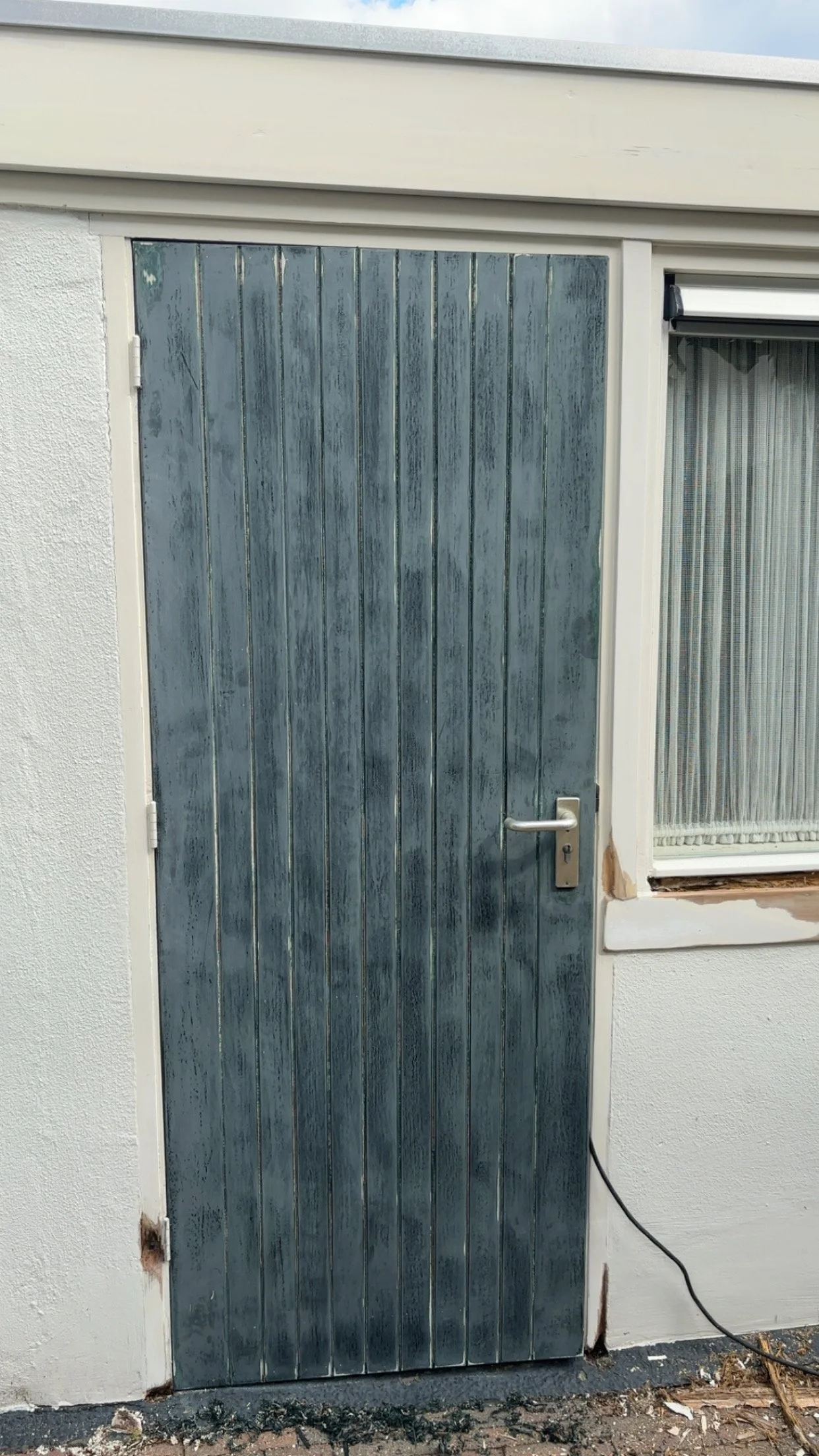 Deur en raam buiten een witte muur, deur van hout met een grijze afwerking, roestplek en waterlek onder het raam, stroomkabel op de grond.