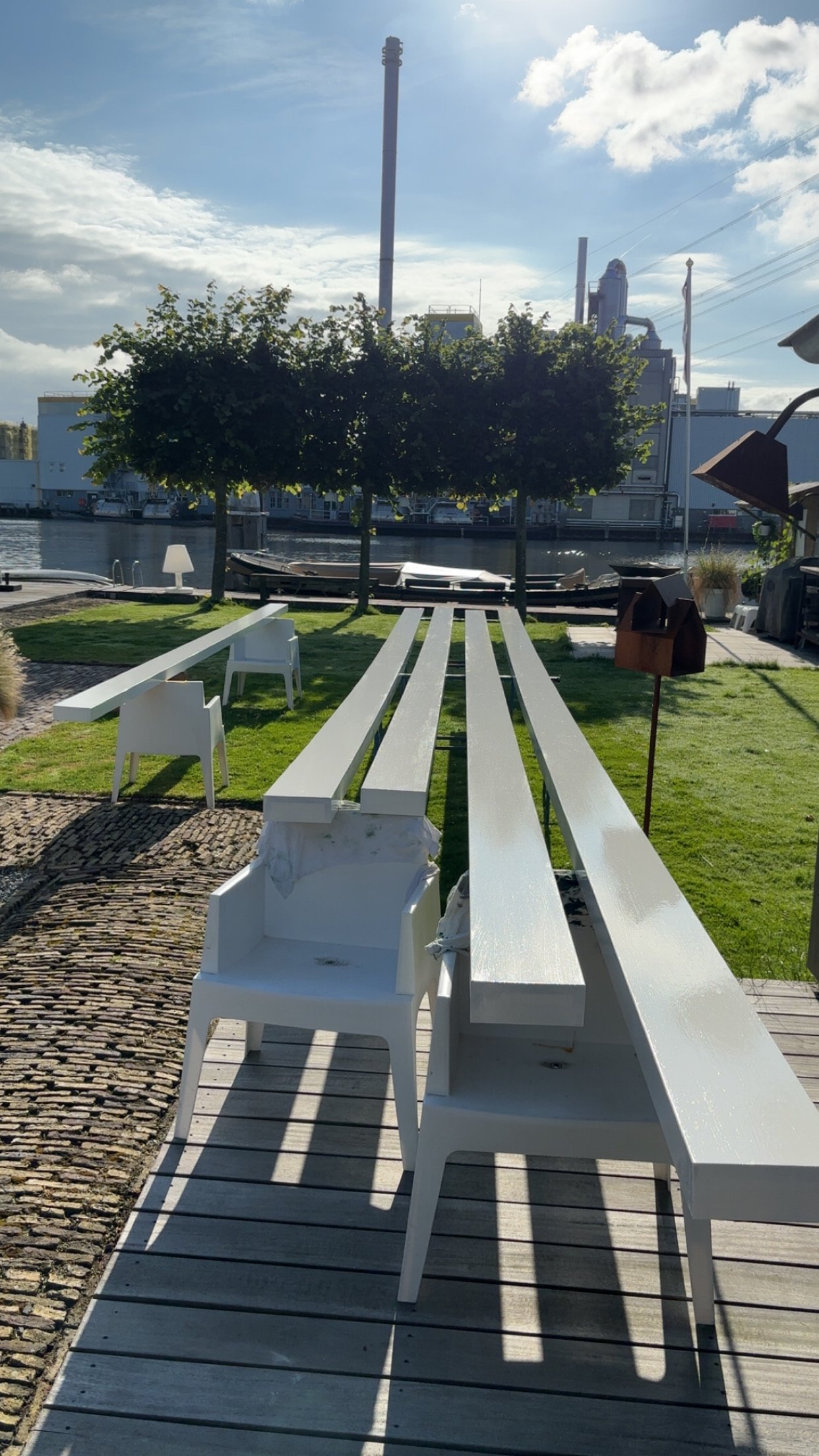 Een buitenterras met witte banken en stoelen, groene gazons, grote bomen, een waterkant en een industrieel gebouw op de achtergrond, onder een bewolkte blauwe lucht.