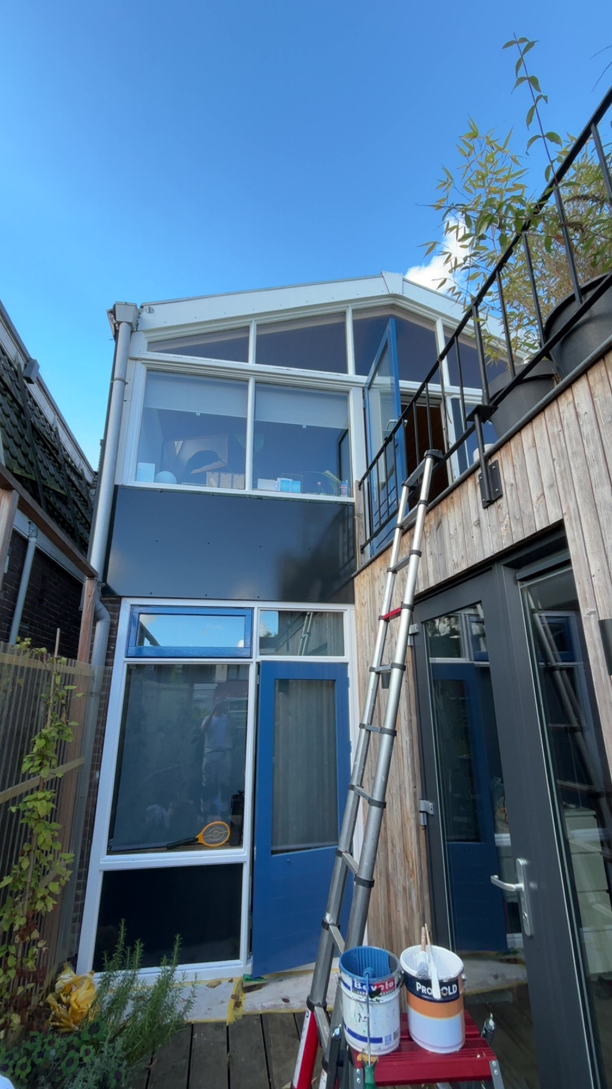 Een modern huis met een houten gevel en grote glazen ramen en deuren. Een ladder en verfemmer staan voor het huis, terwijl een balkon met een zwart hek en planten zichtbaar is.