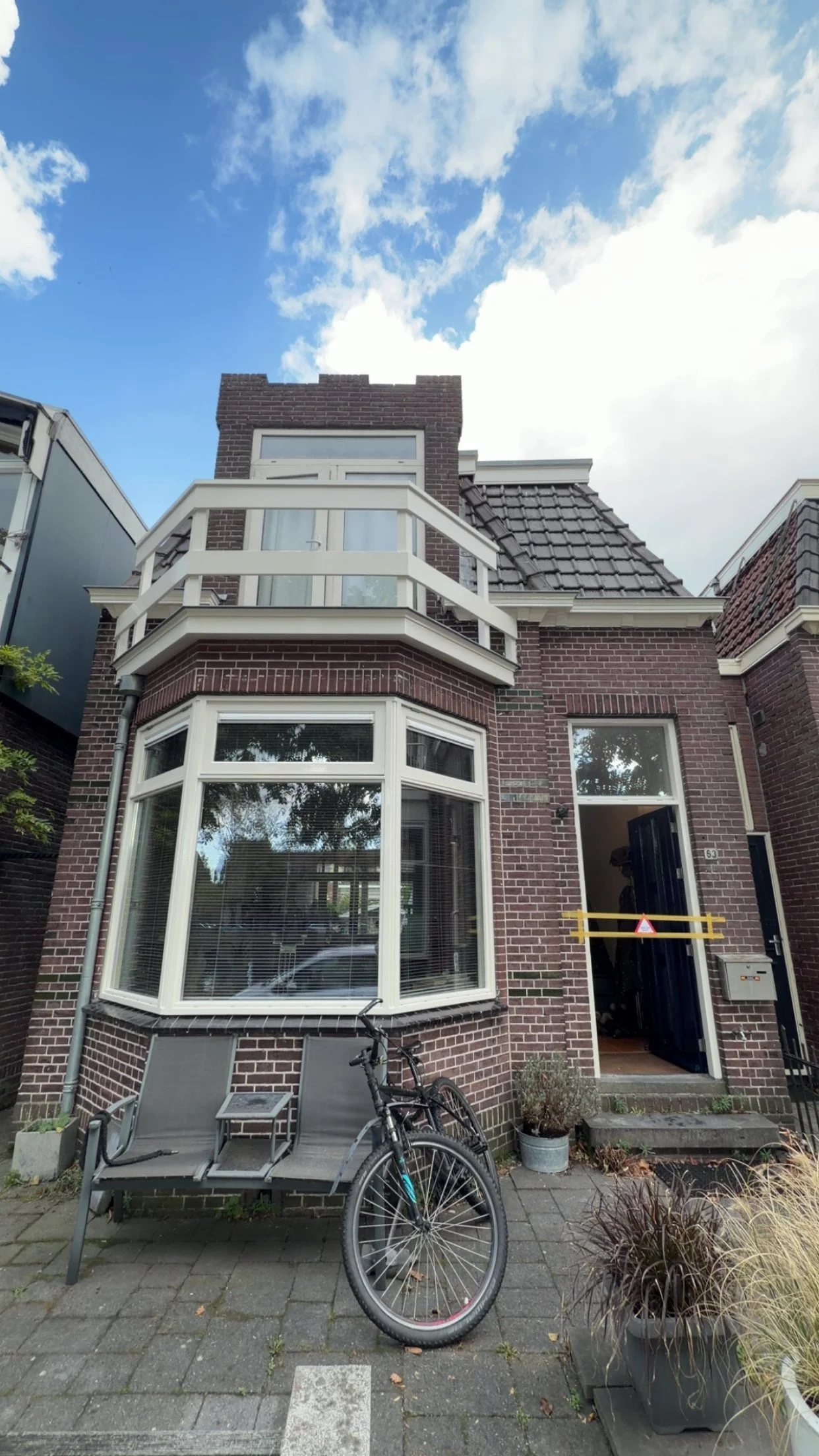Een woning met een bruin bakstenen gevel, hoekramen en een balkon, met een fiets en tuinen voor de ingang.