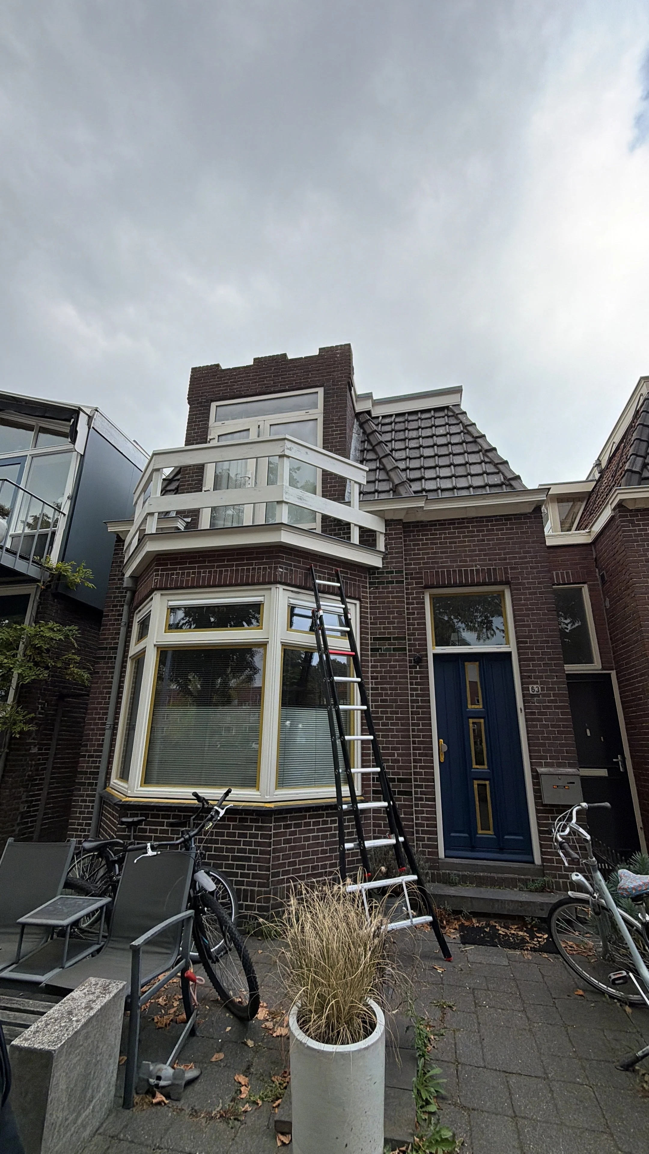 Voorzijde van een rijtjeshuis met een blauwe deur, een grote hoekramen op de begane grond, een balkon op de eerste verdieping, een ladder tegen het huis, fietsen en tuinmeubilair op de voorplaats, en een plant in een grote pot.