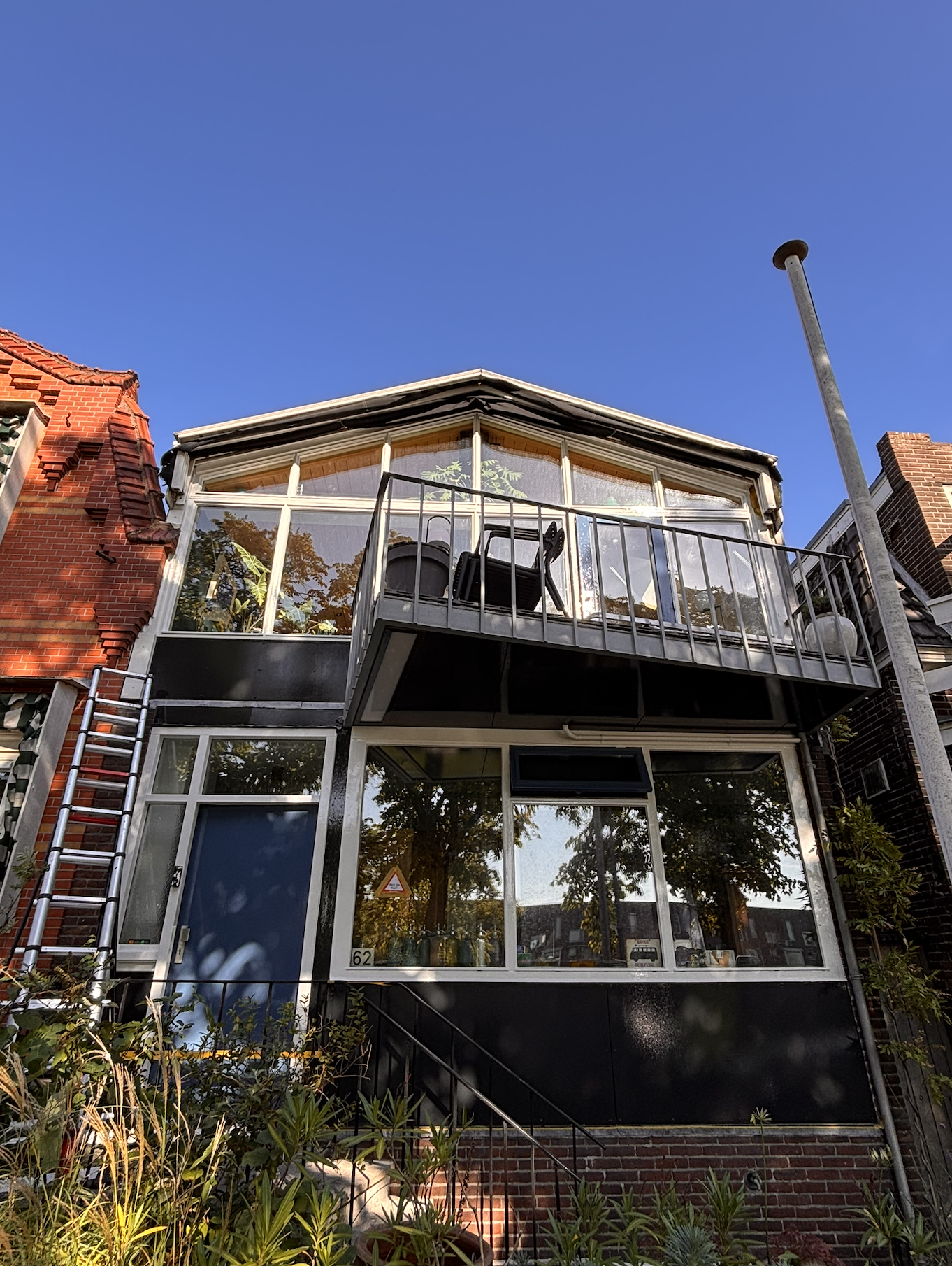 Drie-verdiepings huis met grote ramen, een balkon met tuinmeubilair, en een ladder aan de zijkant, onder een heldere blauwe lucht.