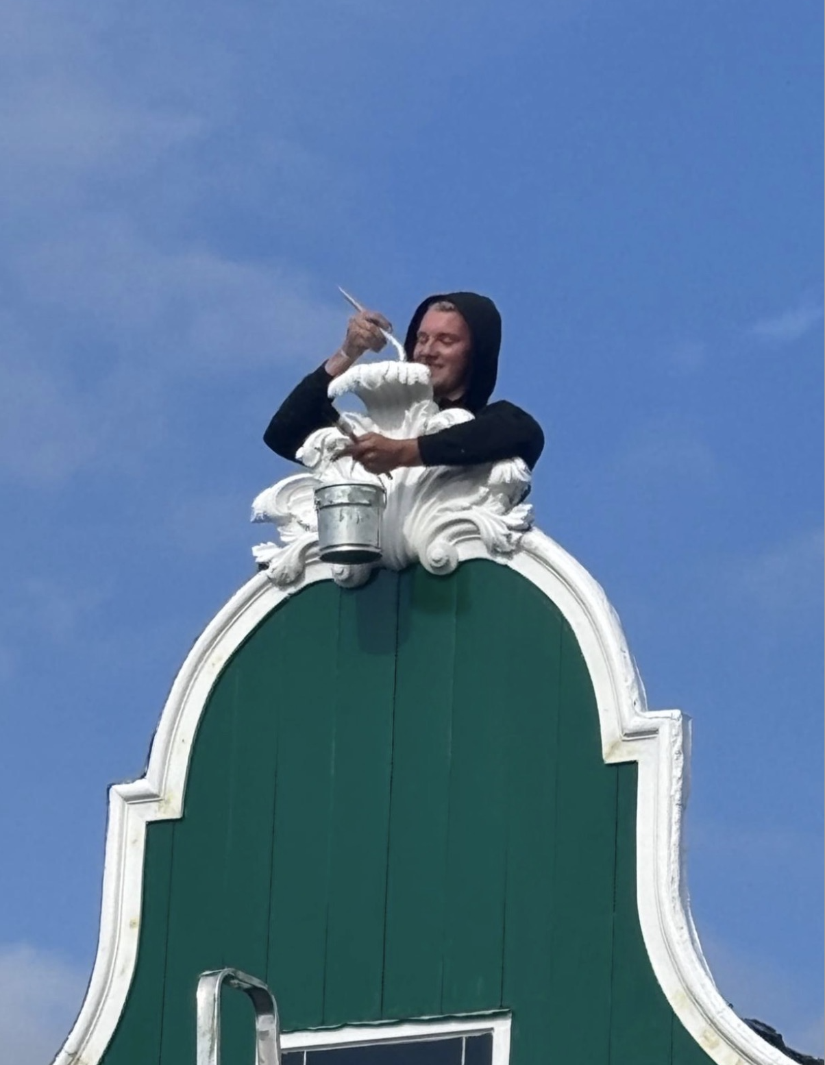 Man with a black hoodie painting a decorative white trim on top of a green building with a blue sky background.