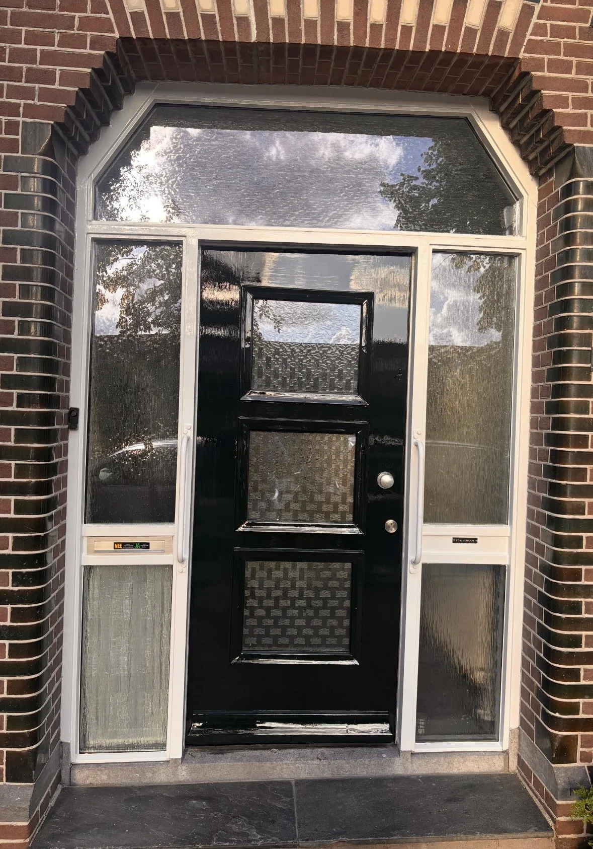 Een zwart voordeur met vier vierkante glas-in-loodpanelen, omgeven door een witte deurkozijn en vaste zijpanelen met vierkante glas-in-loodpanelen. Het huis heeft een bakstenen gevel en er is een boog van bakstenen boven de deur. Het is een dag met e