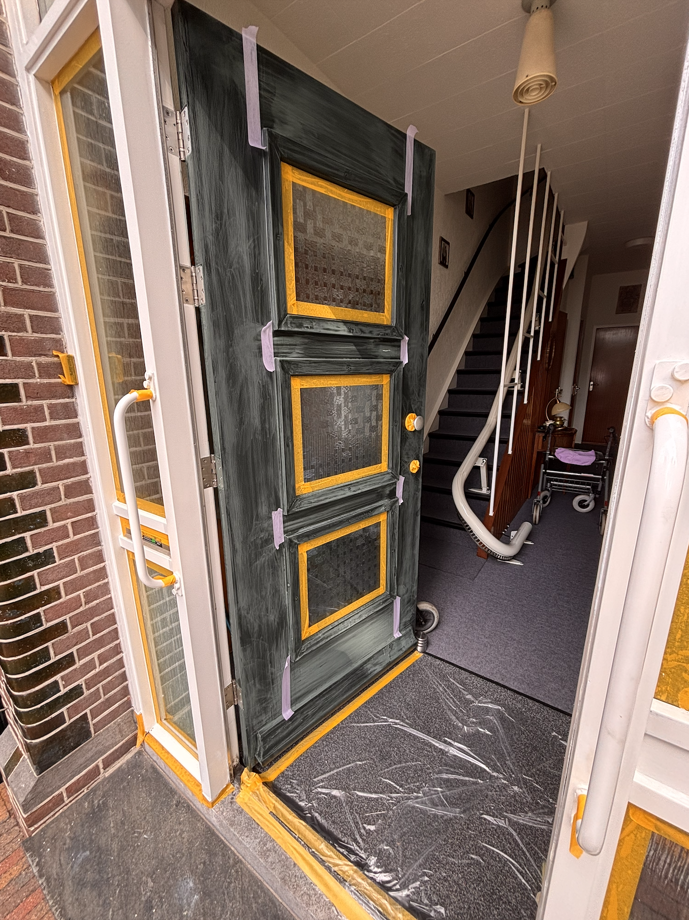 De voordeur van een huis wordt geschilderd, met masking tape om de ramen en randen. De deur is deels geschilderd in een donkere kleur, de muur ernaast bestaat uit bakstenen.