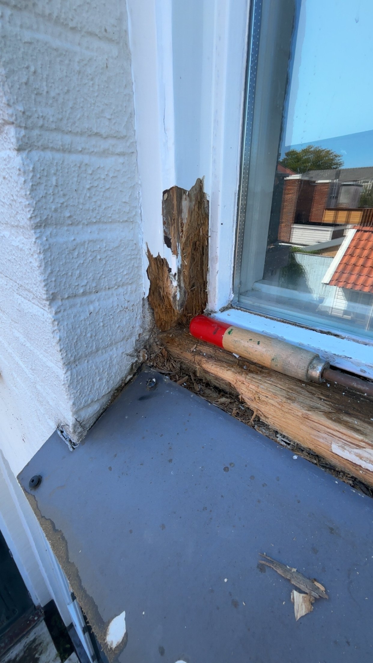 De hoek van een kozijn met beschadigd hout en verfafwijkingen, een verfroller ligt op de houten lijst onder het raam, en er ligt een stuk hout op de betonnen vloer.