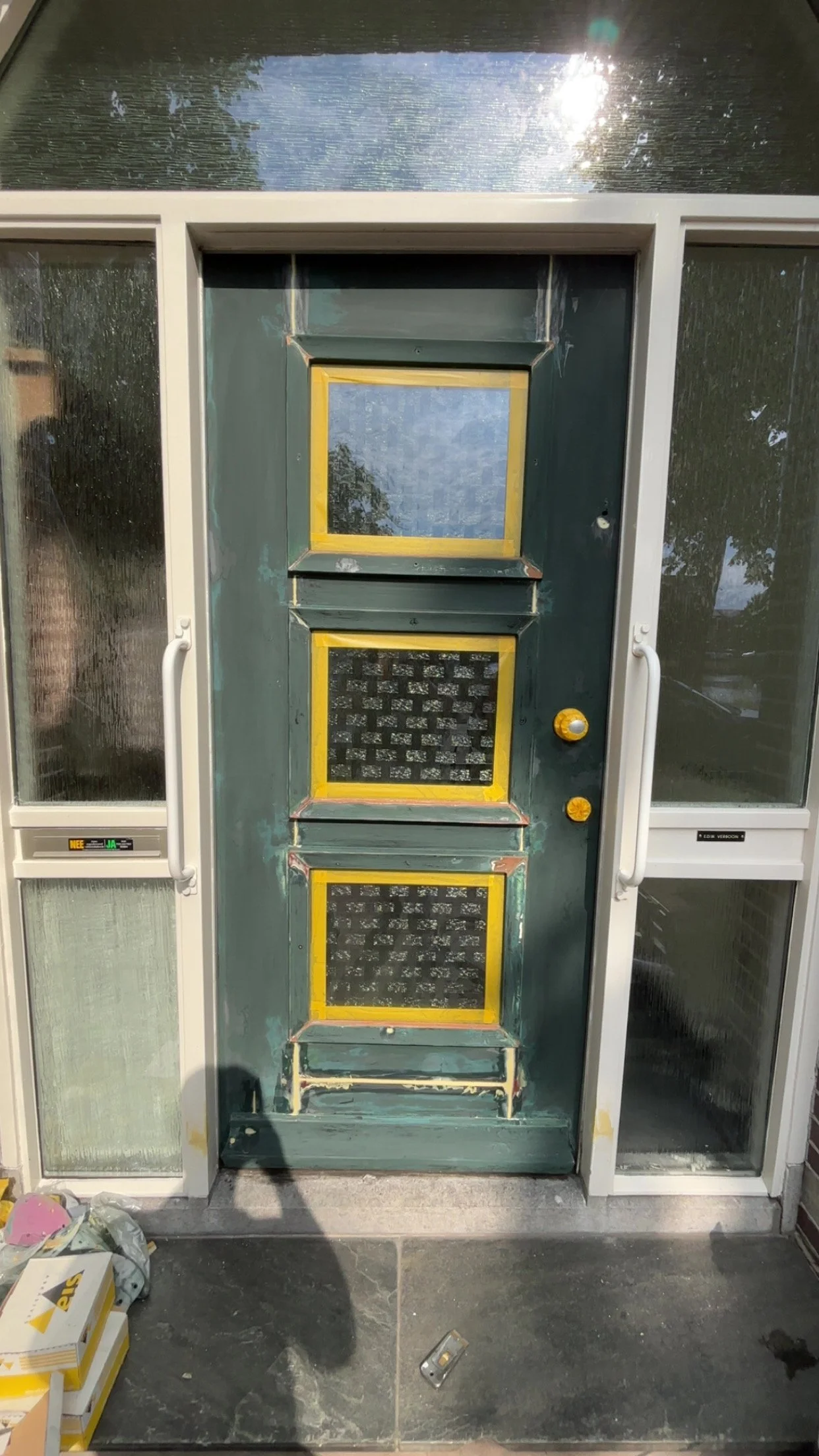 Bouw- of herstelwerkzaamheden aan een groene deur met drie ramen, omringd door andere ramenpanelen, met verf- en gereedschap op de vloer.