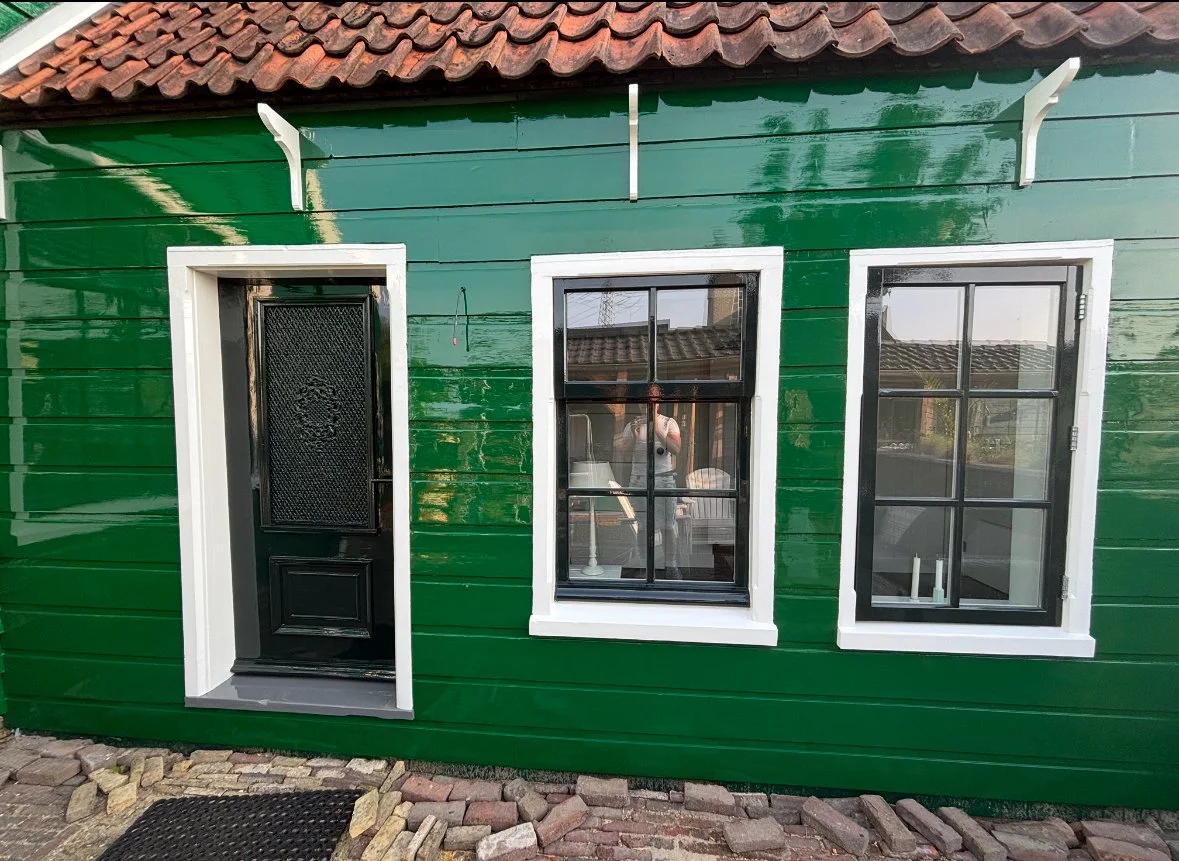 Groene houten gevel met deur en twee ramen, rode dakpannen en stenen oprit.