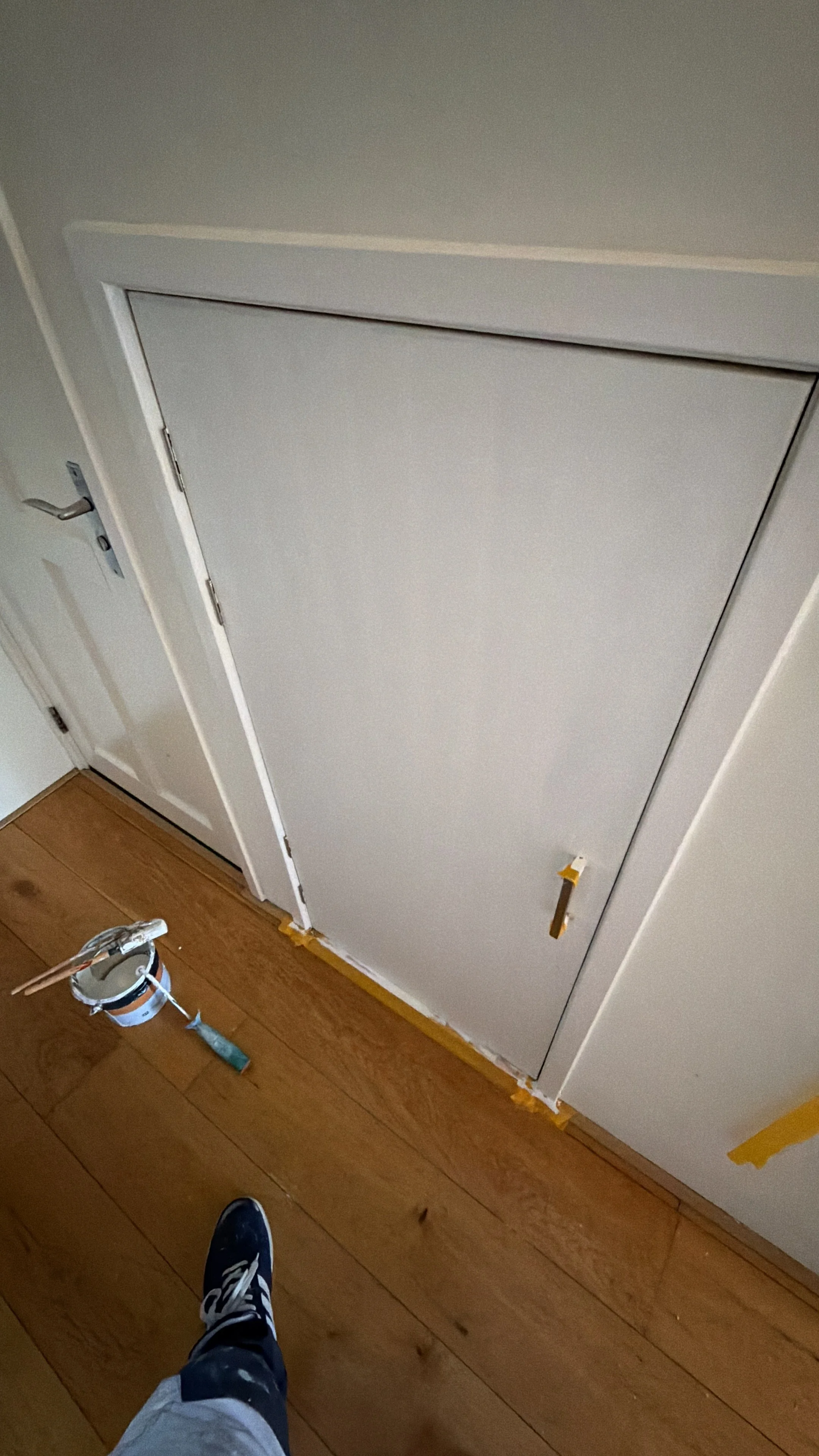 Binnenkant van een gesloten, wit paneeldeur met een schroefgat en een handvat. De deur bevindt zich in een witte muur. Op de houten vloer staan een verfbakje en een verfroller. Een persoon draagt sneakers en lichte broek.