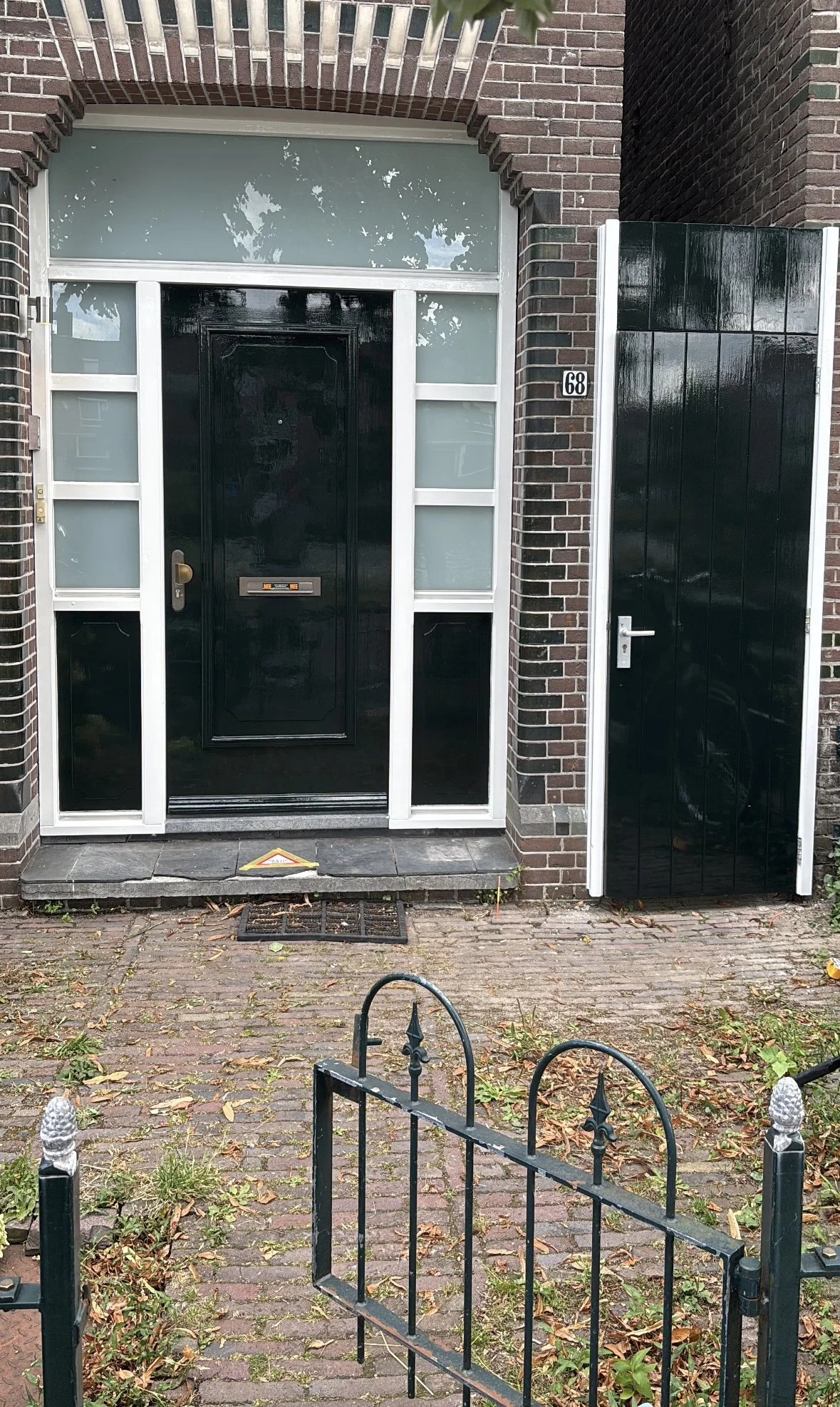 De afbeelding toont de voordeur van een huis met een zwart deurkozijn en een zwarte deur. Er is een kleine oprit met een metalen hek voor de ingang, en een huisnummer '68' aan de muur links van de deur. Er liggen bladeren op de bestrating.