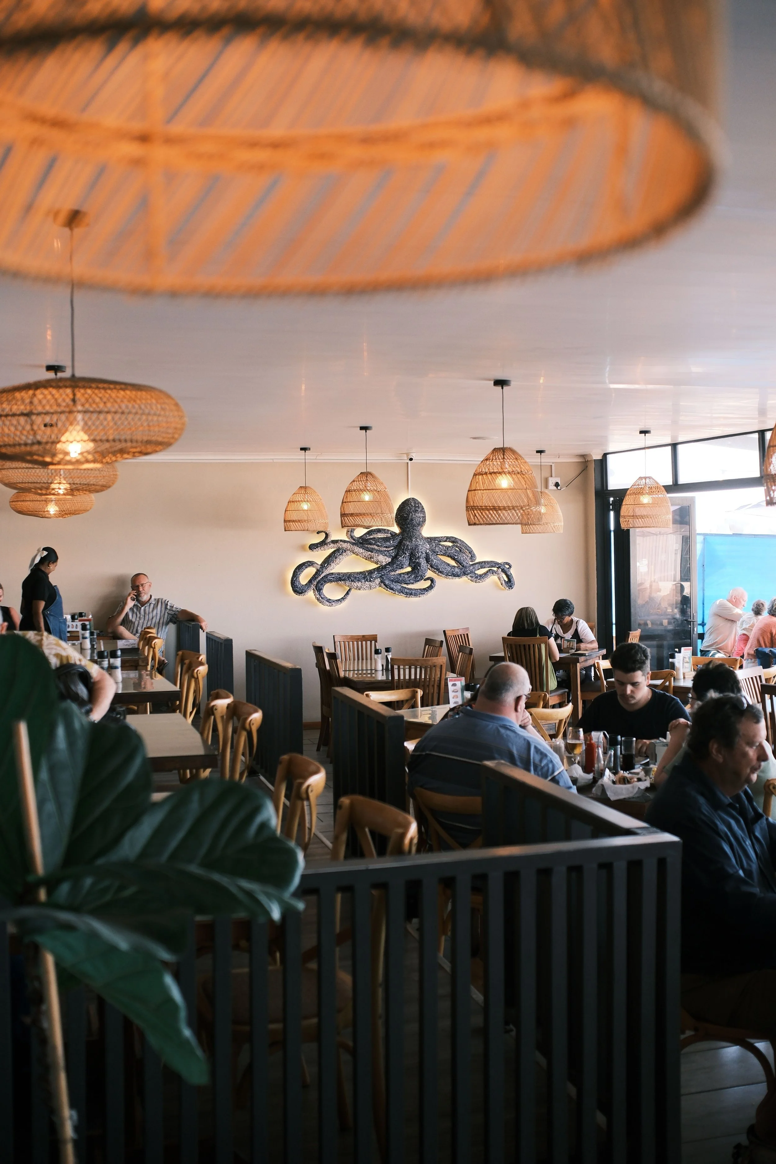 Interior of a restaurant with wicker pendant lights, a black octopus wall sculpture, and people dining at tables.