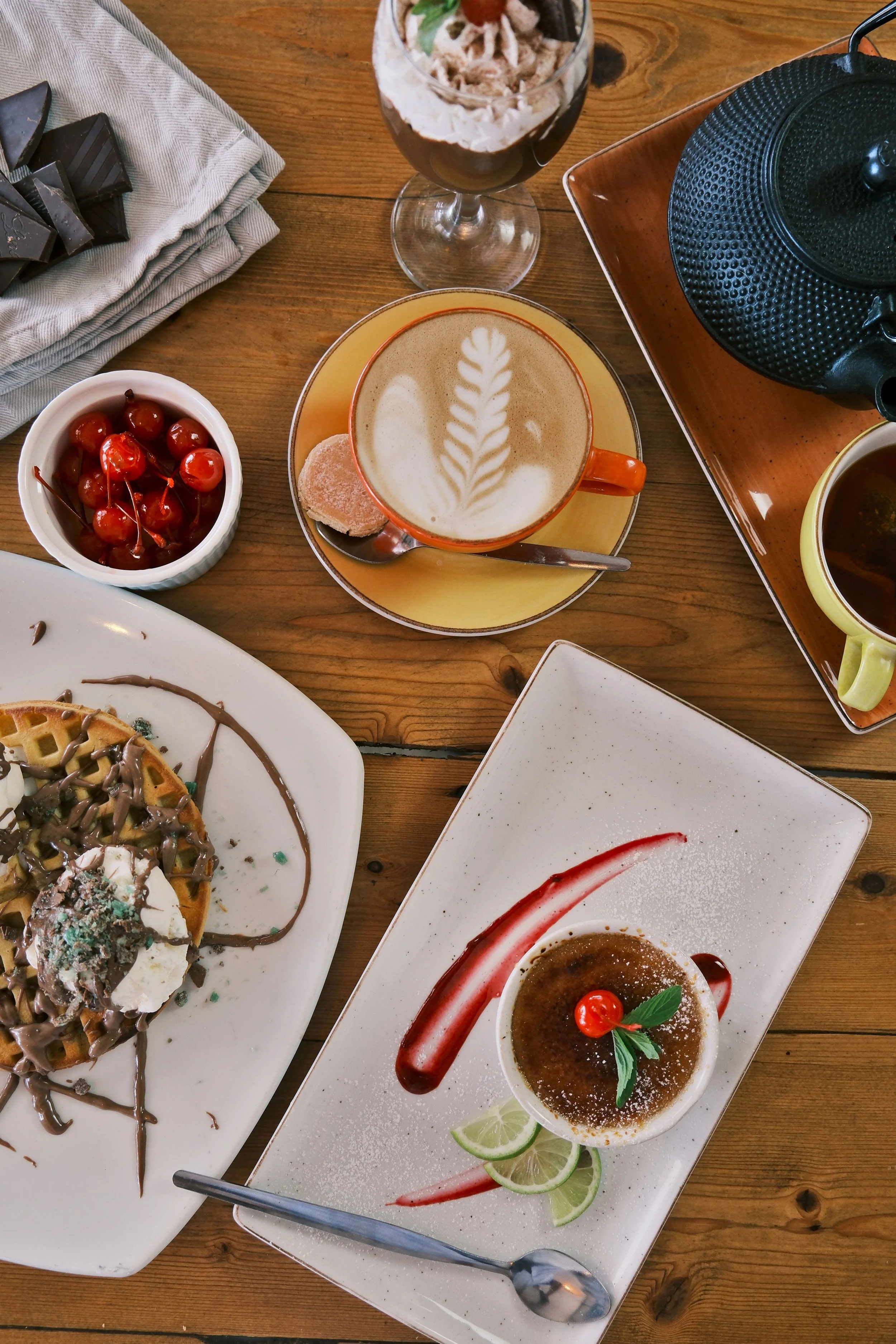 Assorted desserts, coffee drinks, cherries, and chocolates on a wooden table, with a teapot and cups visible.