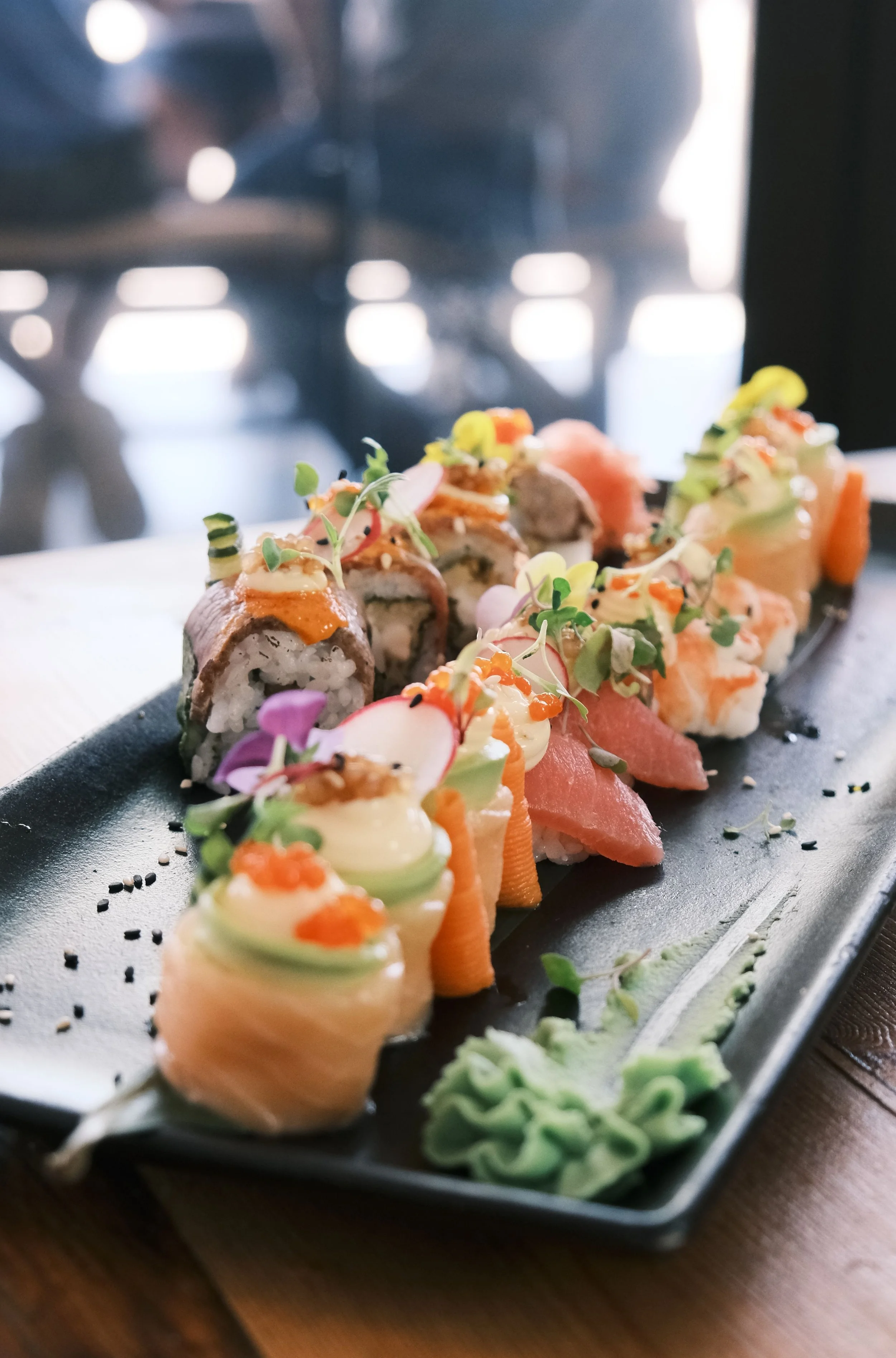 Assorted sushi rolls with various toppings, garnished with microgreens and wasabi on a black rectangular plate.