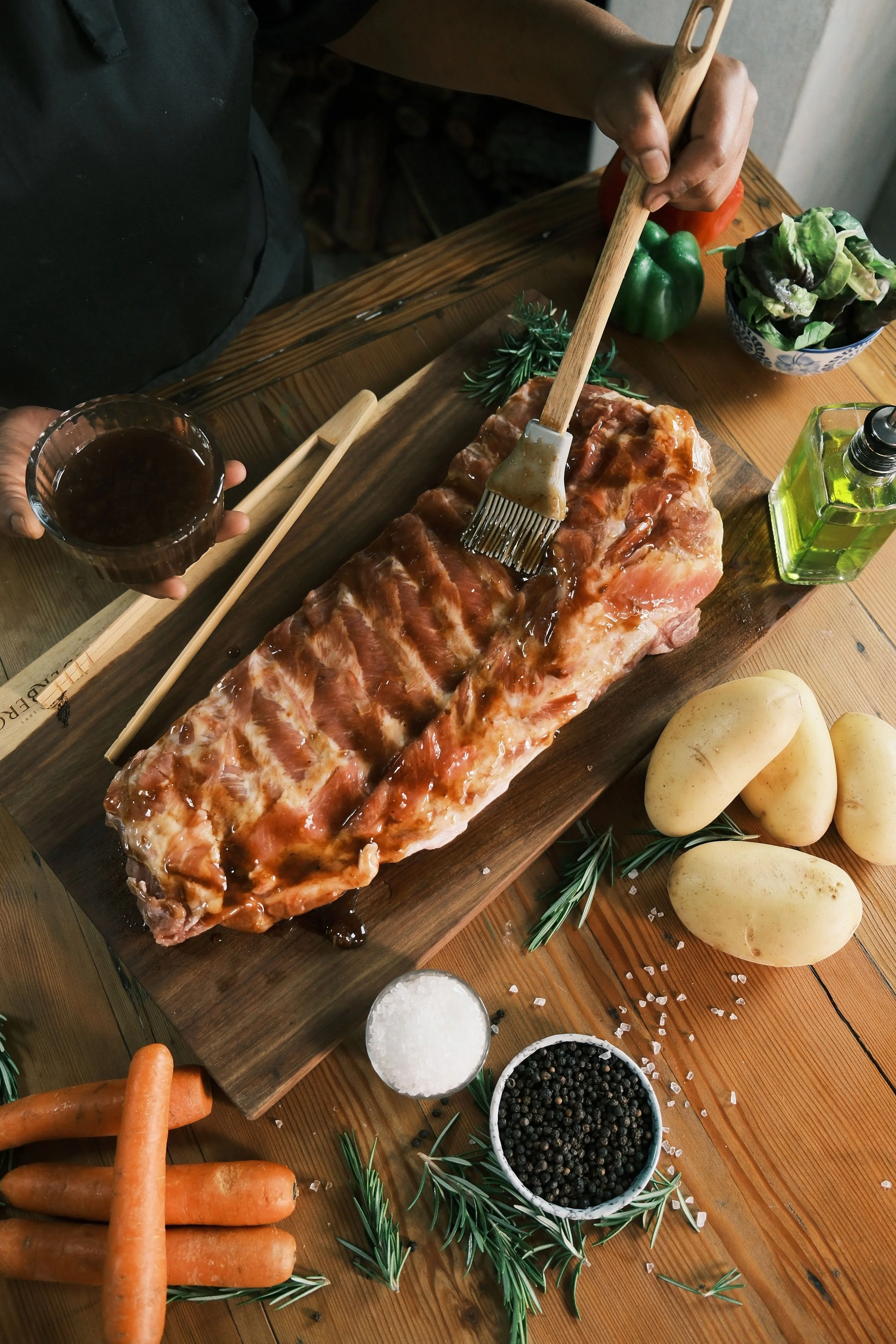 Person basting a raw rack of ribs with barbecue sauce on a wooden table surrounded by carrots, potatoes, herbs, salt, pepper, a bowl of greens, and a bottle of oil.