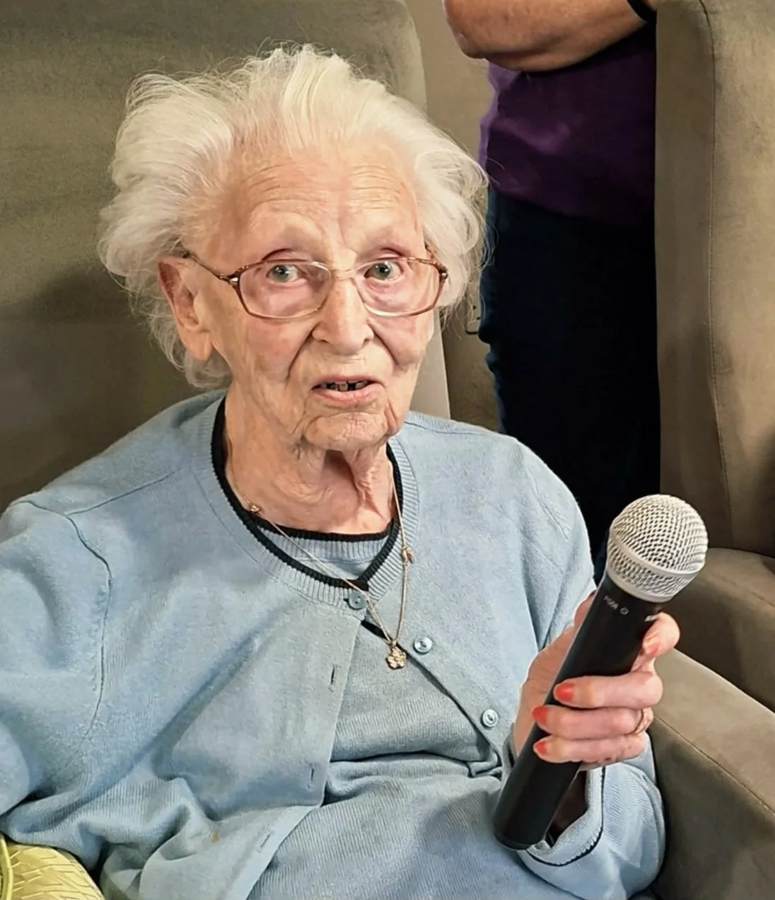 An elderly woman with white hair, glasses, and a light blue sweater, holding a microphone, sitting on a beige chair, with a person standing behind her.