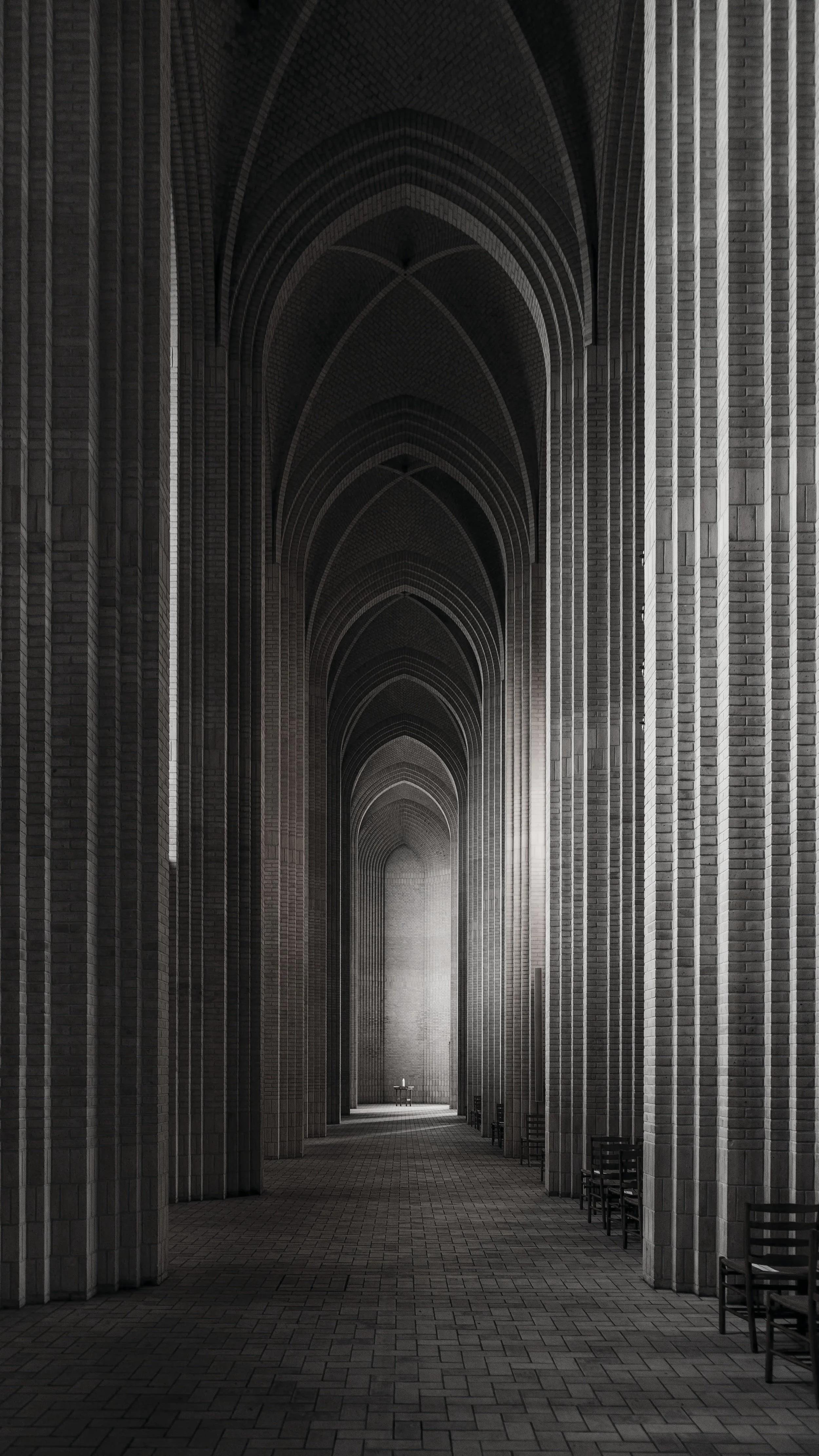 Interior view of a dark, cathedral-like hallway with high arched ceilings, brick walls, and chairs lined along the sides, leading to a lit area with a cross in the distance.
